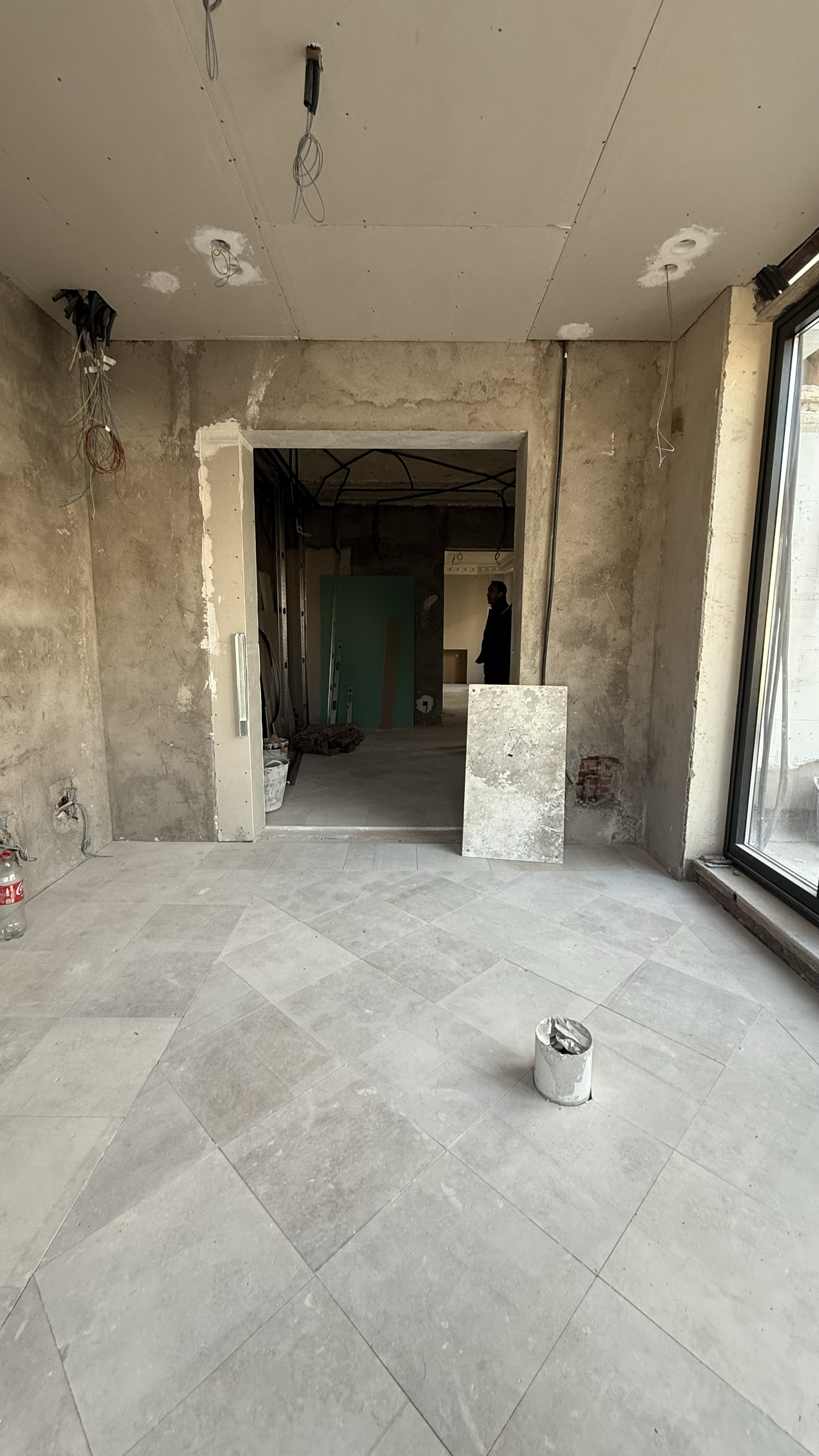 Interior of a room under renovation, with exposed drywall, construction materials, and partially finished tiled floor, and a man standing in the background.