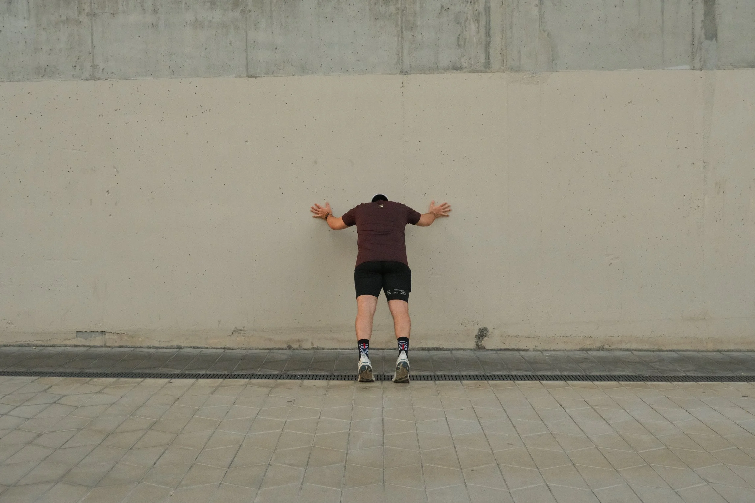 A person kneeling with their hands and forehead pressed against a concrete wall in an indoor or outdoor space with tiled pavement.