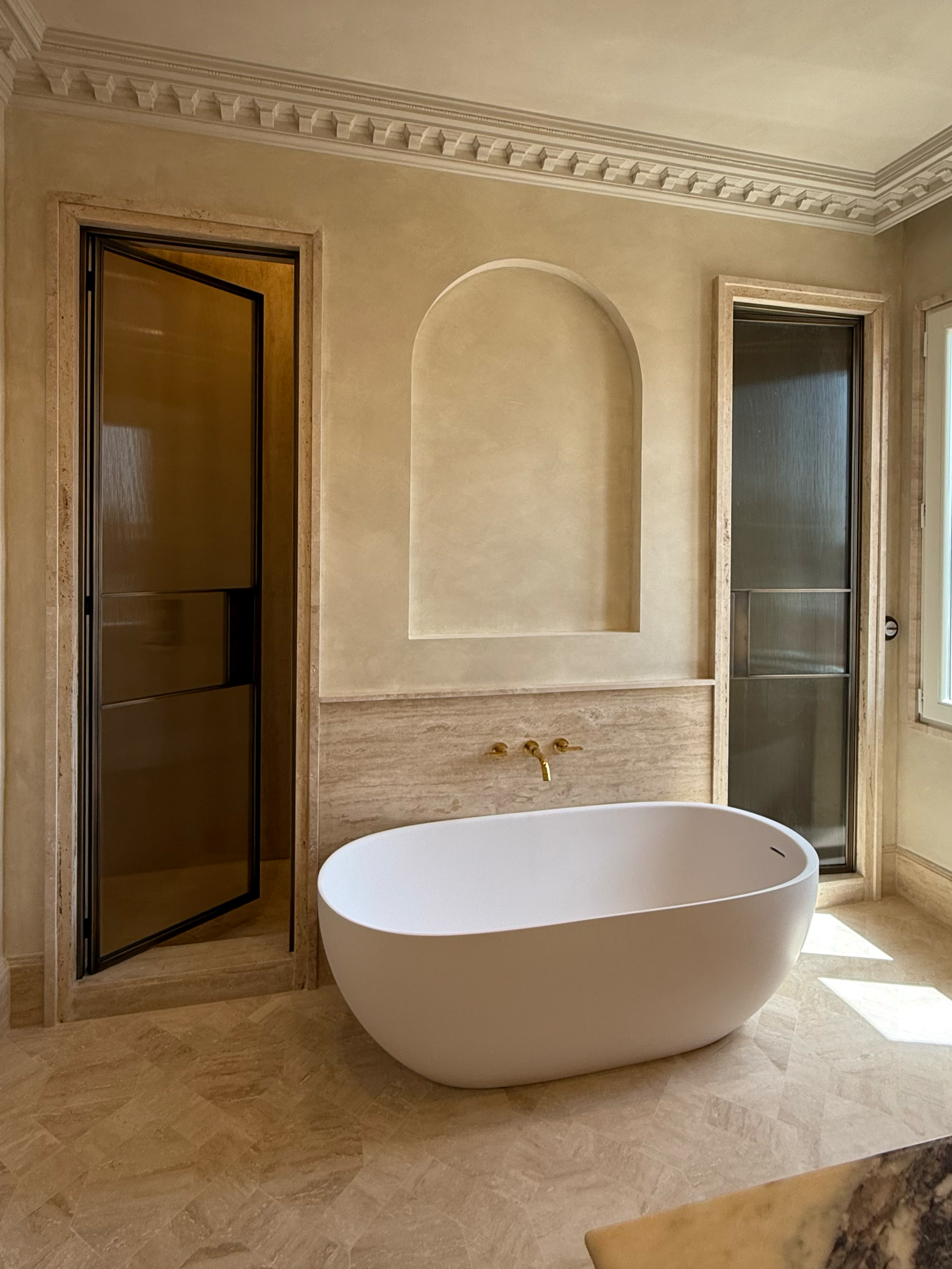 Modern bathroom with a freestanding bathtub, two enclosed showers with glass doors, and gold fixtures, all featuring beige and cream marble finishes.