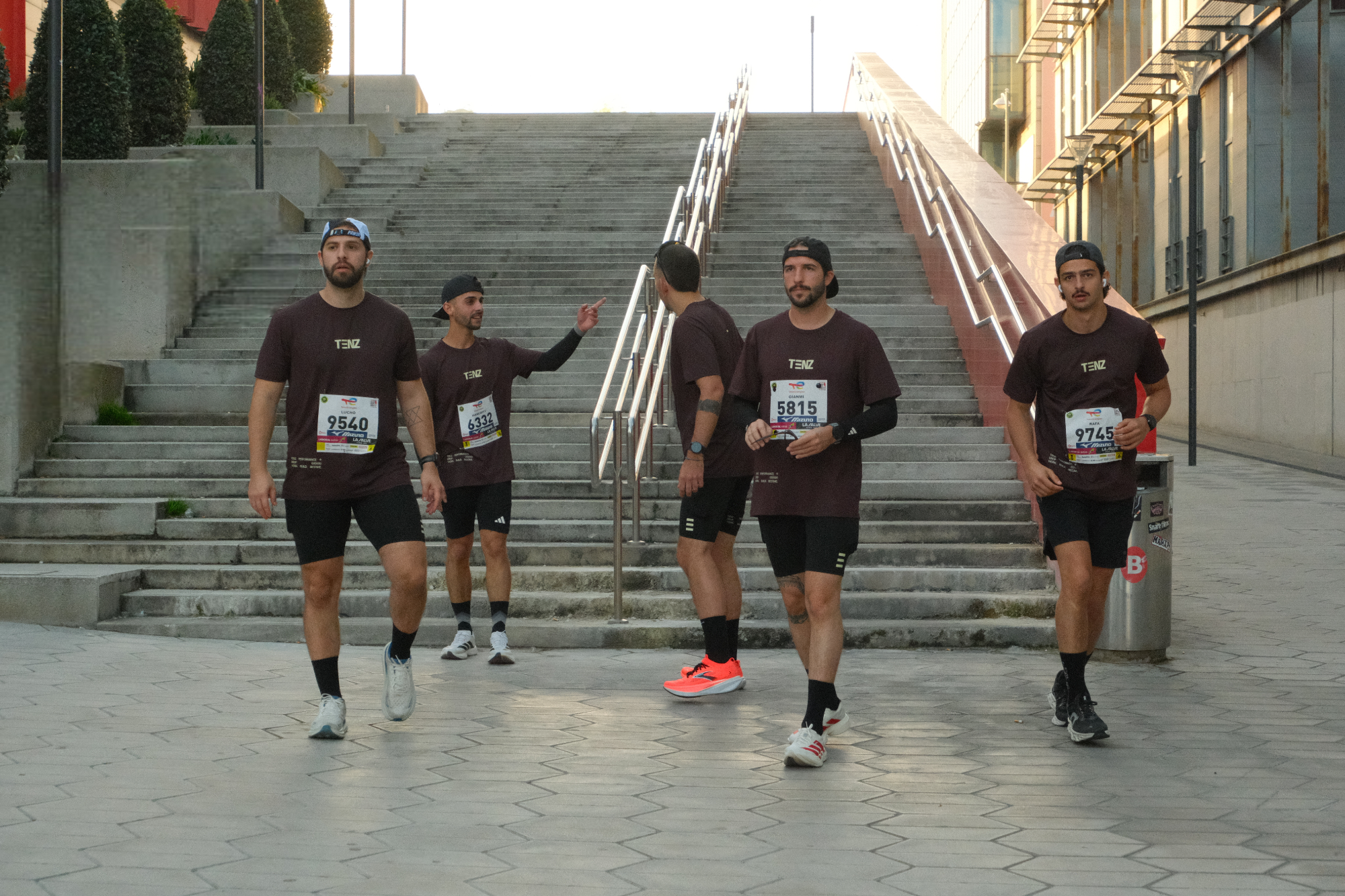 Five men in matching dark purple t-shirts and black shorts participating in a running event, walking away from a staircase in an urban setting with modern buildings.