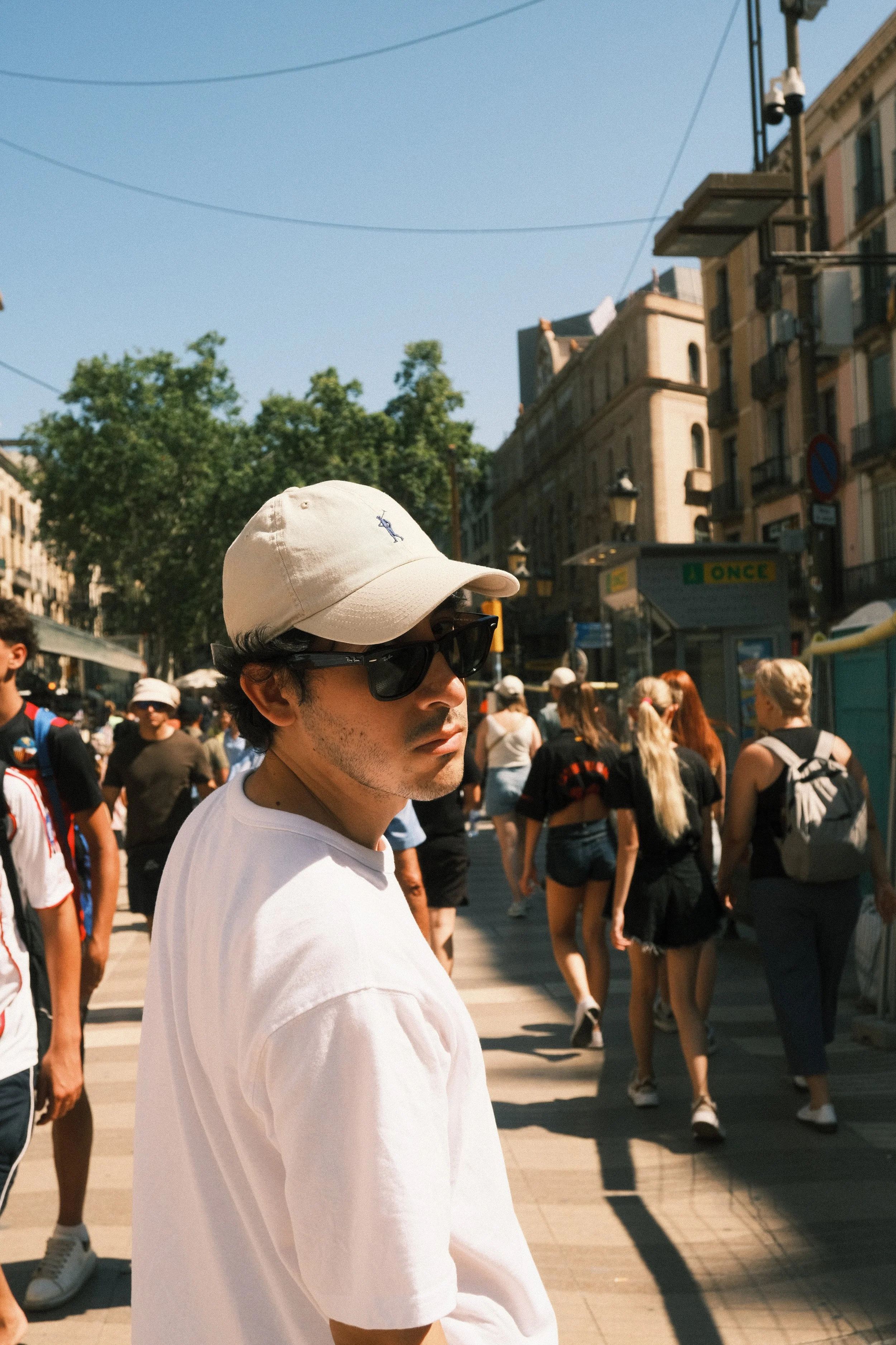 A man wearing sunglasses, a beige cap, and a white shirt walking in a busy city street on a sunny day, with many people around and old buildings in the background.