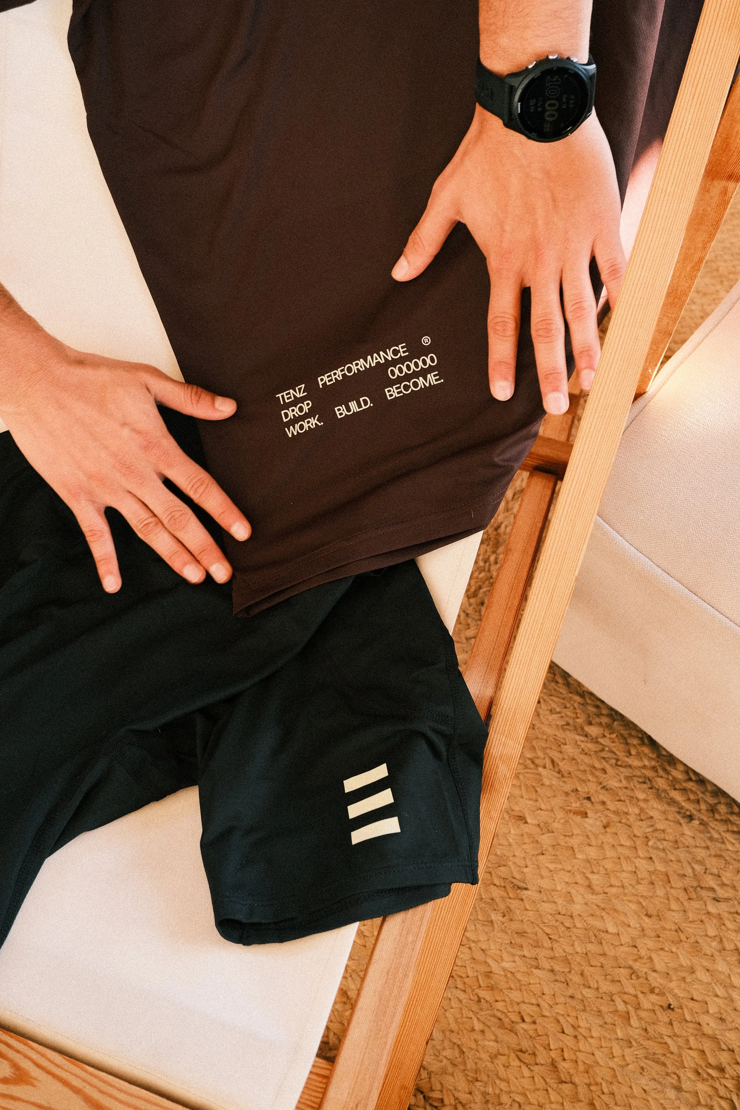 Person sitting on a bench wearing a black sports watch and black athletic shorts with three white stripes, holding a brown Tenz Performance bag with white text, near a beige chair and woven carpet.