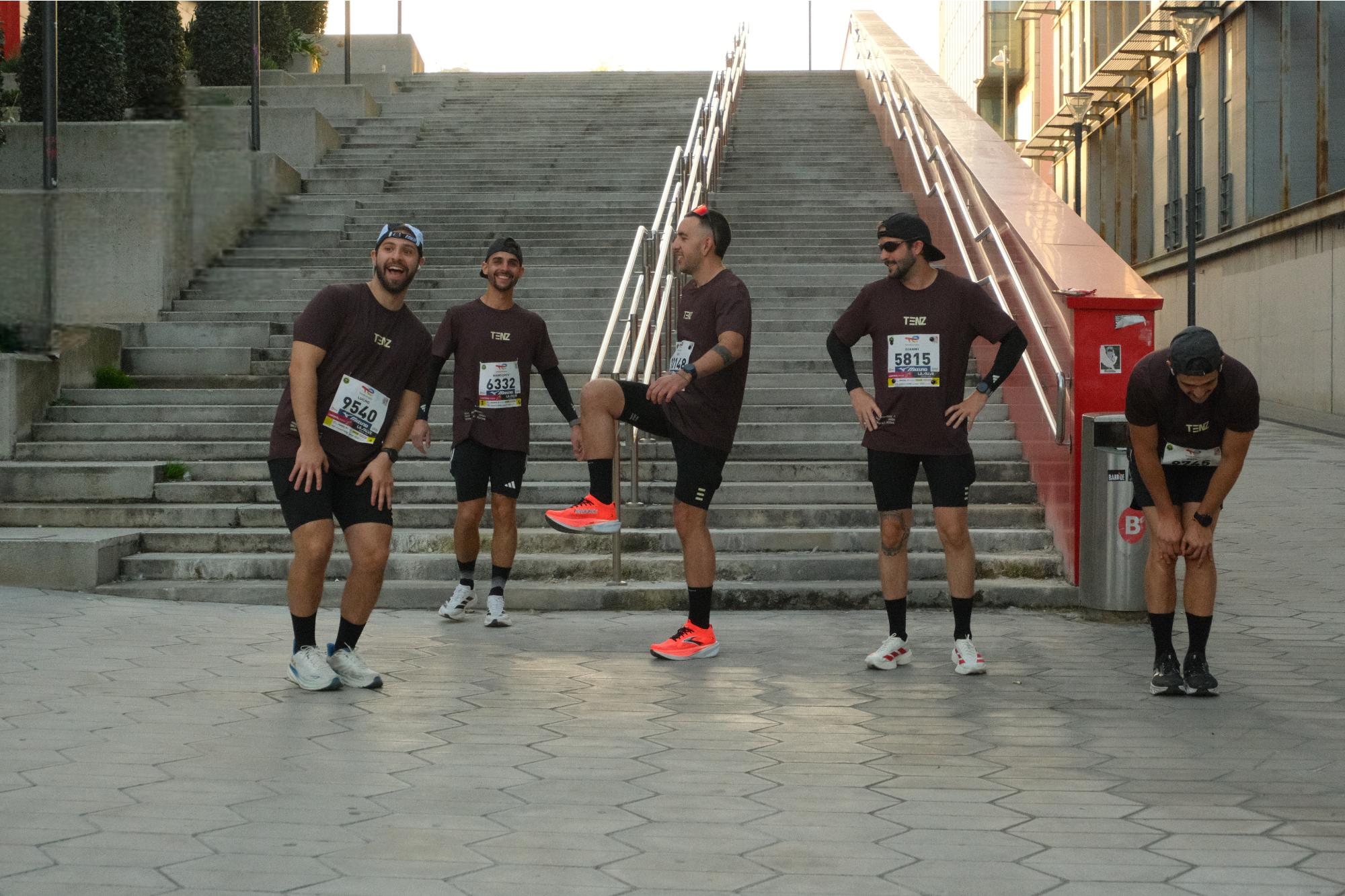 Group of five men in athletic wear taking a break after running, standing on a city street in front of stairs and a red trash bin.