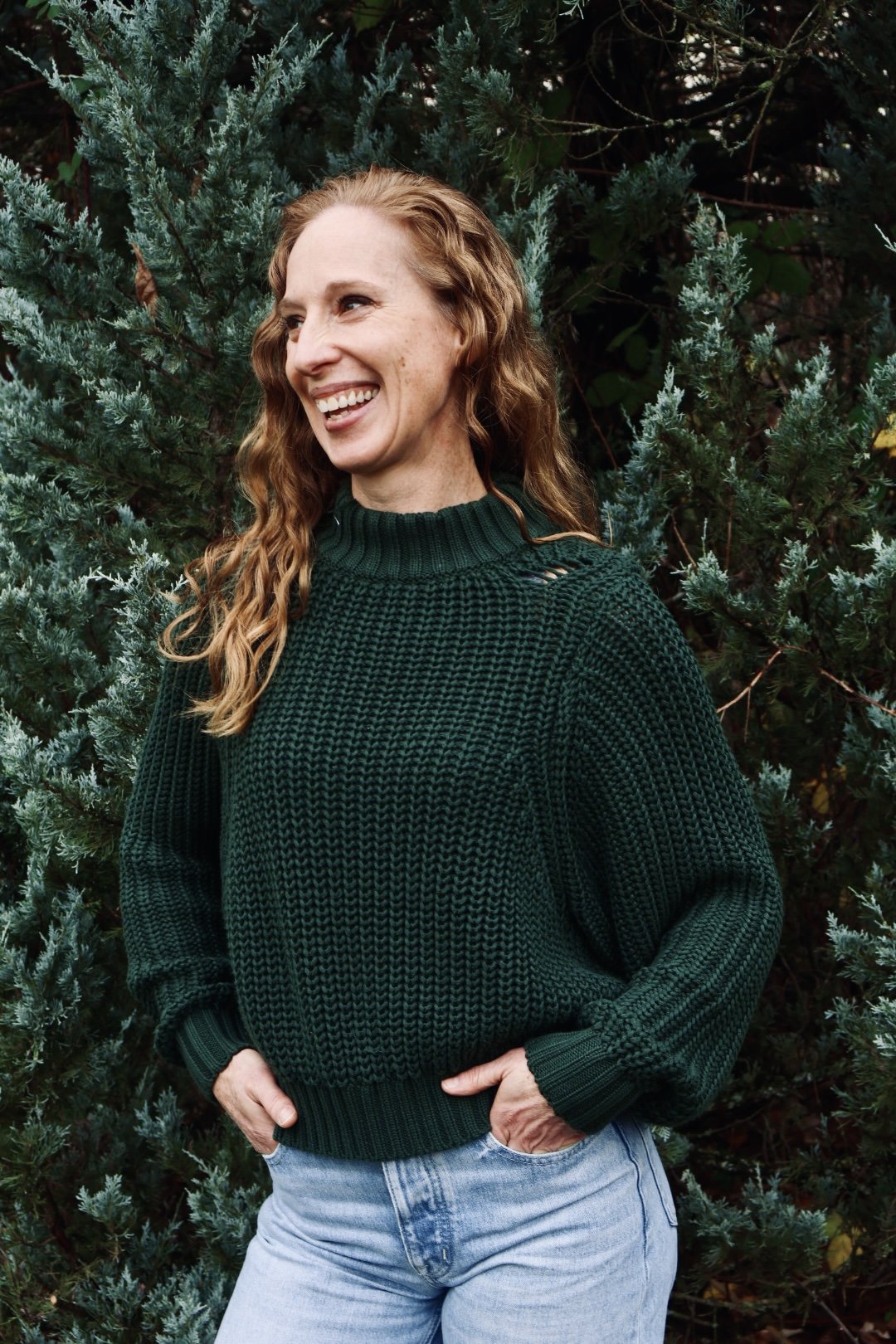 A woman with curly, red hair smiling and looking to her left, standing outdoors in front of green foliage, wearing a dark green knitted sweater and light blue jeans.