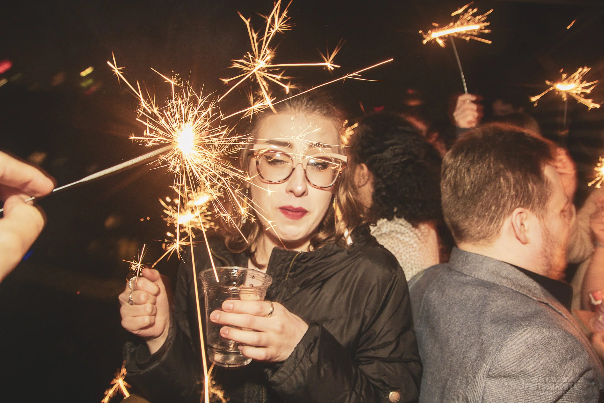 A woman holding a sparkler and a clear cup at a celebration or party with other people in the background.