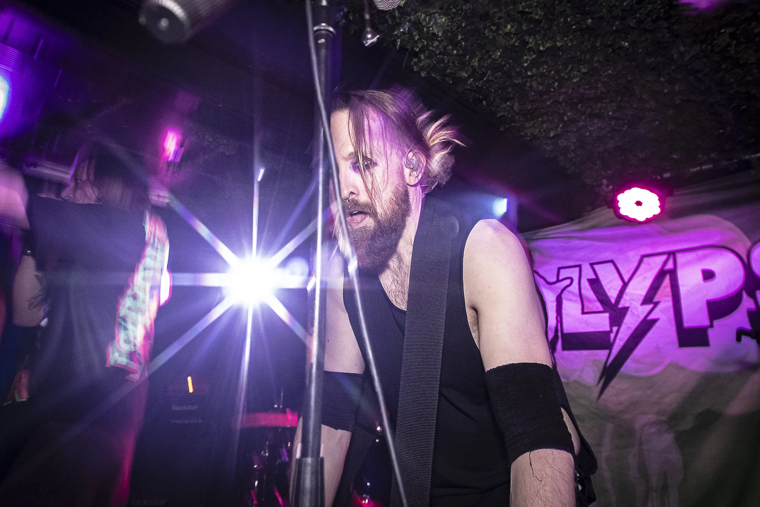 A man with a beard and long hair playing a musical instrument in a dimly lit venue with colorful lighting and a banner with lightning bolt symbols in the background.