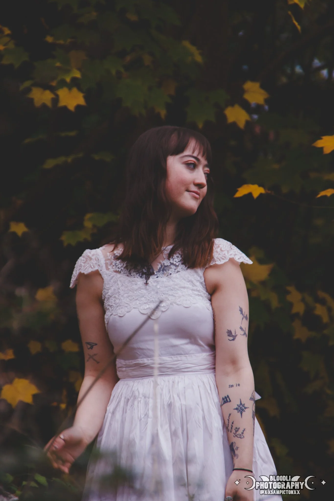 A young woman with shoulder-length brown hair and tattoos on her arms, wearing a white lace dress, standing outdoors with a background of autumn leaves.