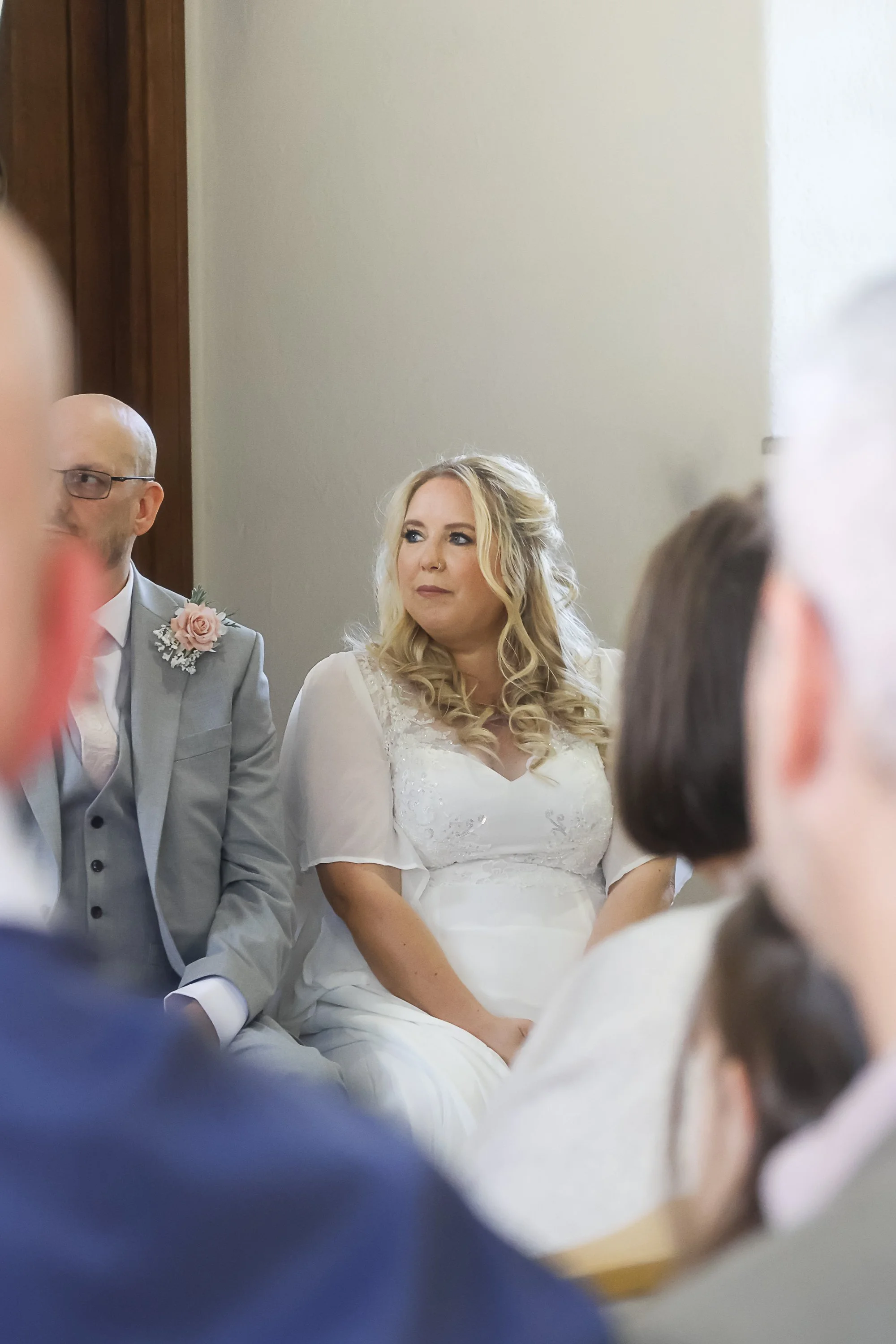 A woman with blonde hair wearing a white dress sitting in a wedding ceremony; a man in a grey suit with a pink boutonniere is sitting next to her, and other guests are partially visible surrounding them.
