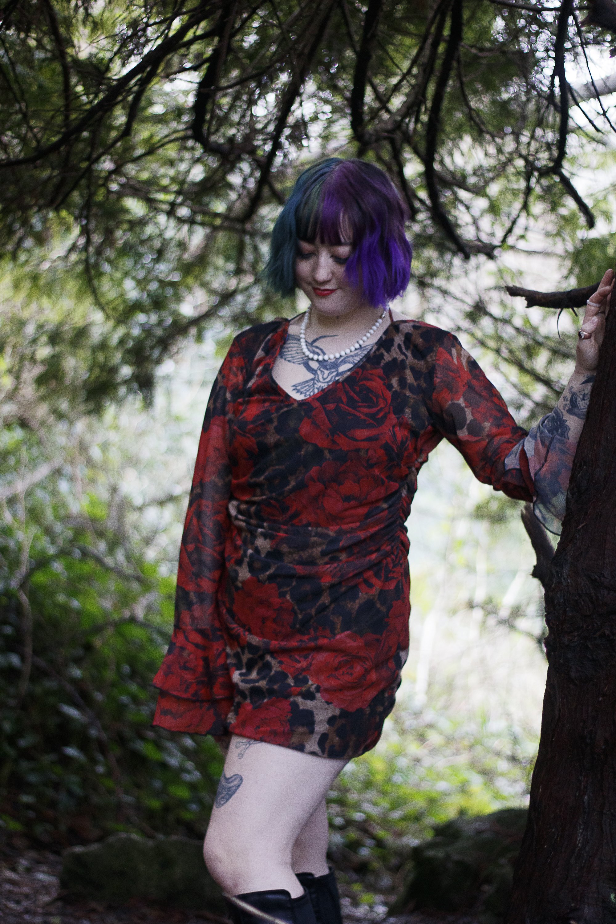 A woman with purple and blue hair, wearing a red floral dress, standing in a wooded area with her hand on a tree branch, smiling softly.