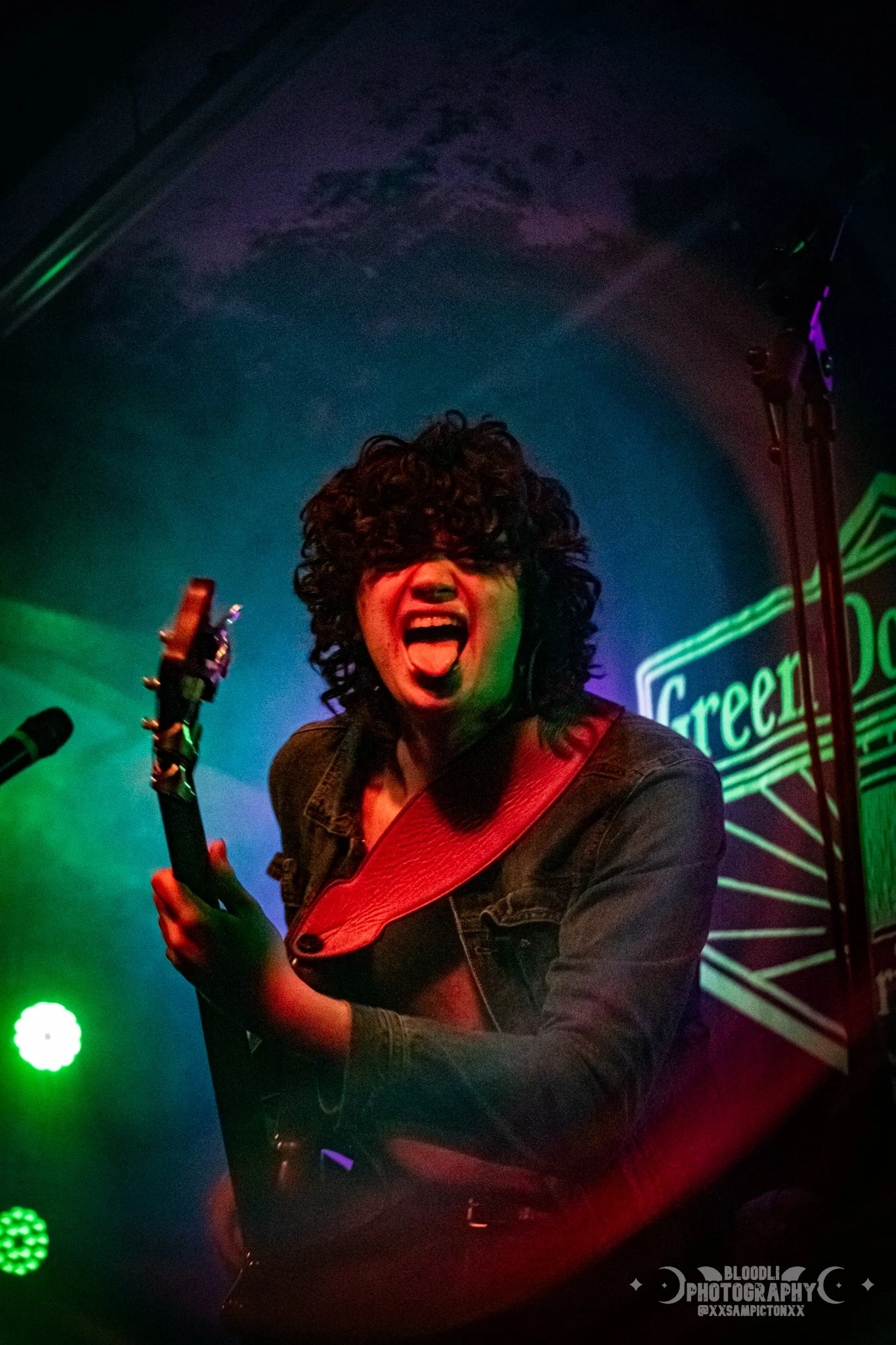 A woman with curly black hair passionately singing or screaming while playing an electric guitar on stage with colorful lighting and a green neon sign that reads 'Greet'.