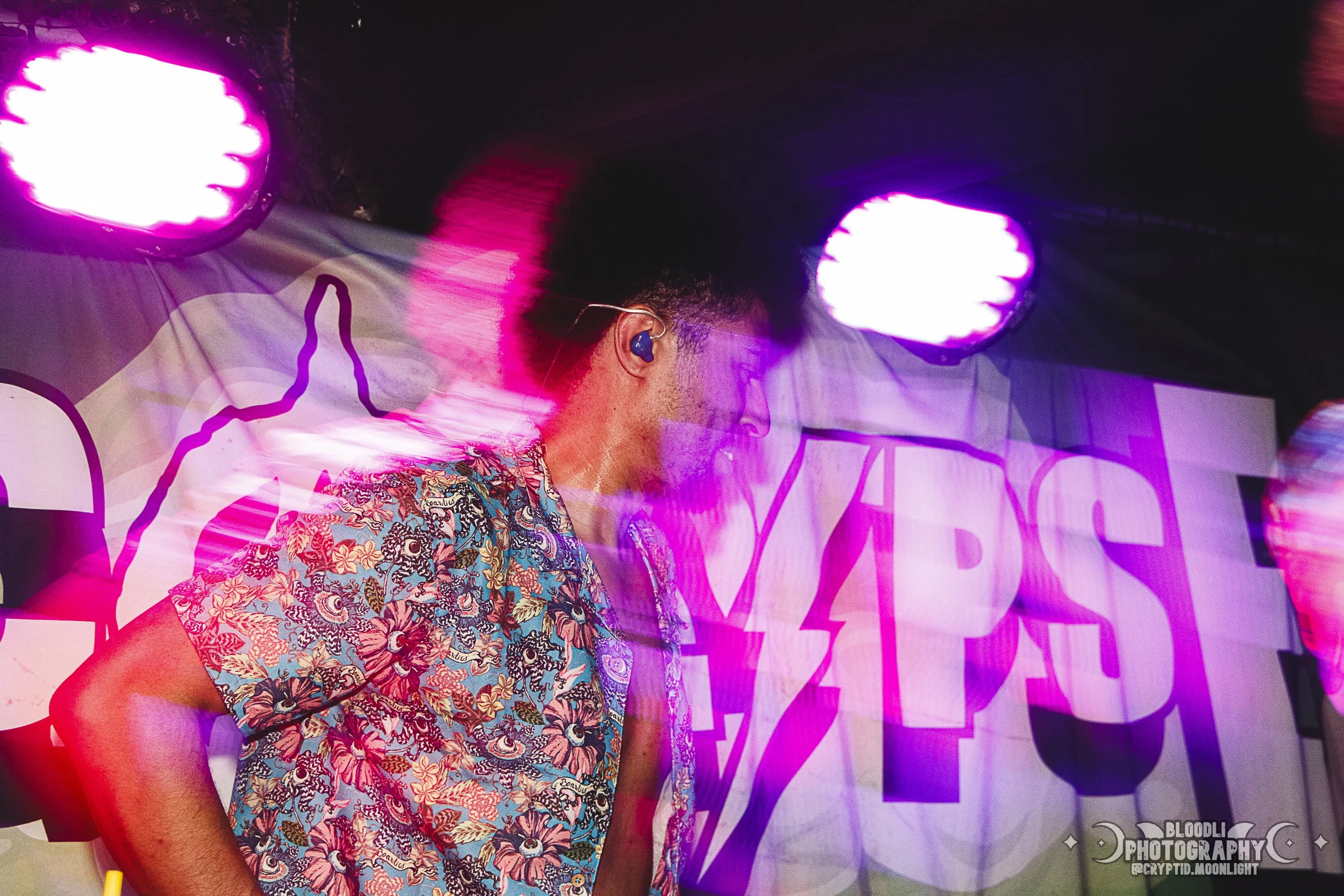 A person on stage with colorful lights, wearing a floral shirt, and wearing in-ear monitors, with a backdrop featuring bold graphics and text.