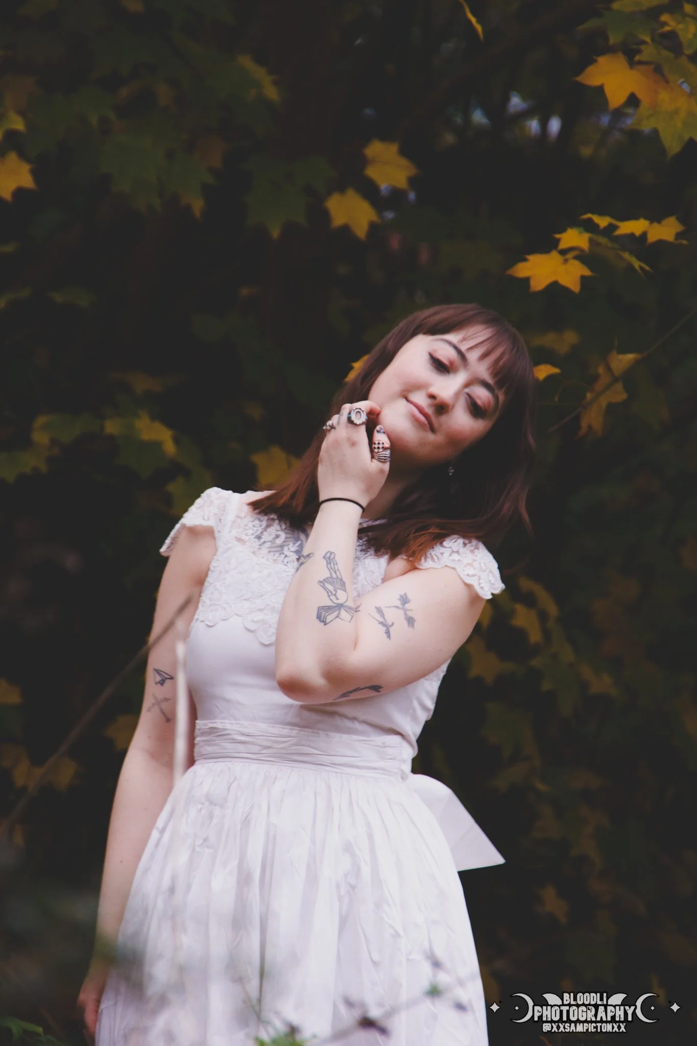A woman in a white dress with lace detail on the shoulder, standing outdoors with yellow leaves and dark foliage in the background. She has tattoos on her arms, rings on her fingers, and is posing with her hand near her face, smiling softly.