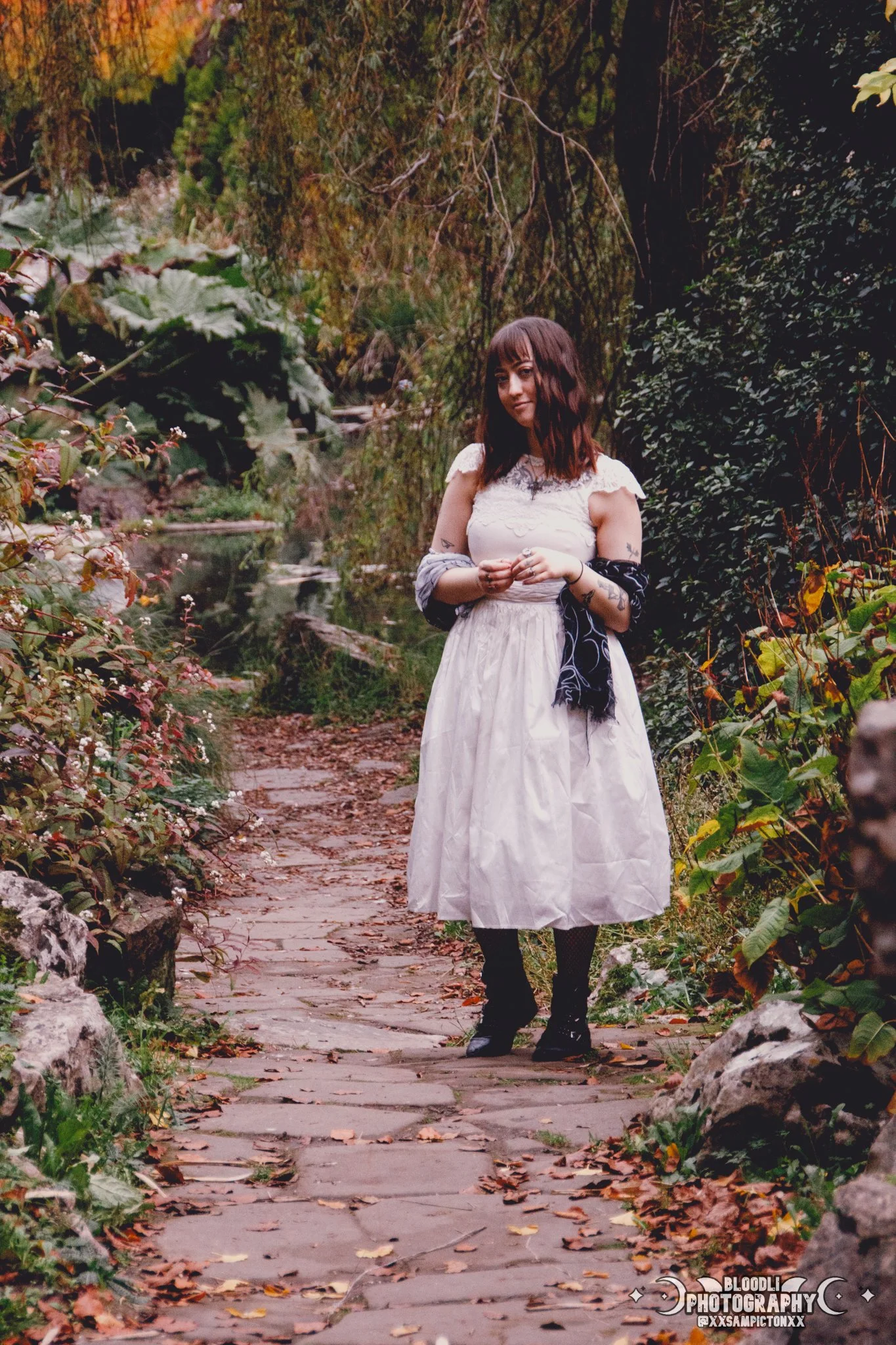 A woman with dark hair in a white dress standing on a stone pathway in a lush garden with autumn leaves and foliage.
