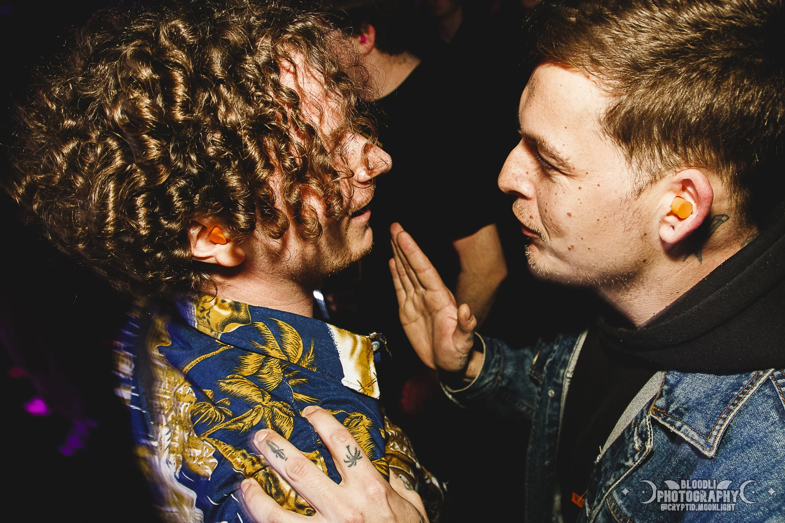 Two men facing each other closely, with one touching the other's face in a tense interaction, both wearing ear plugs.