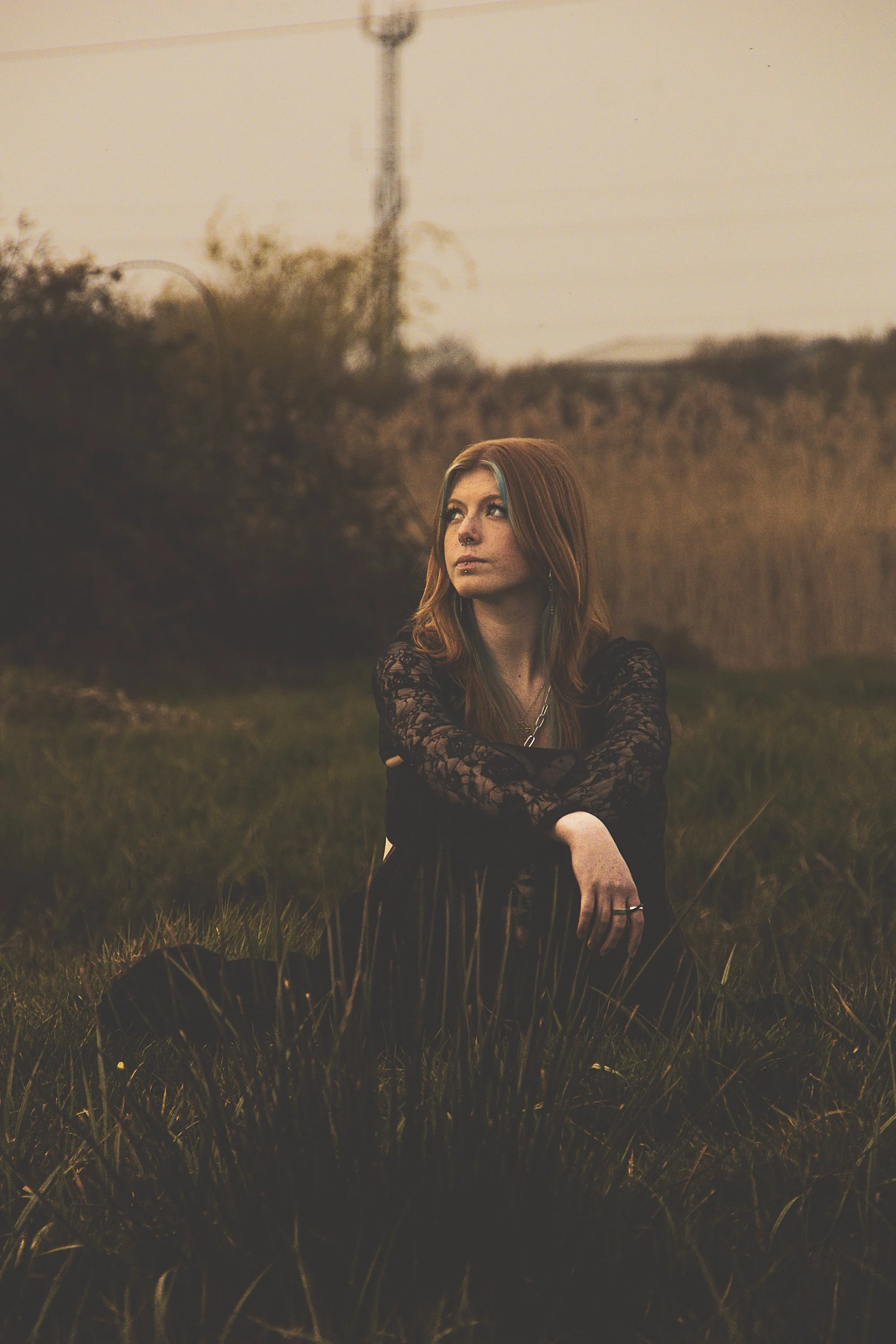 A young woman with red hair and a septum piercing sitting in a field at sunset, wearing a black lace top and black pants.