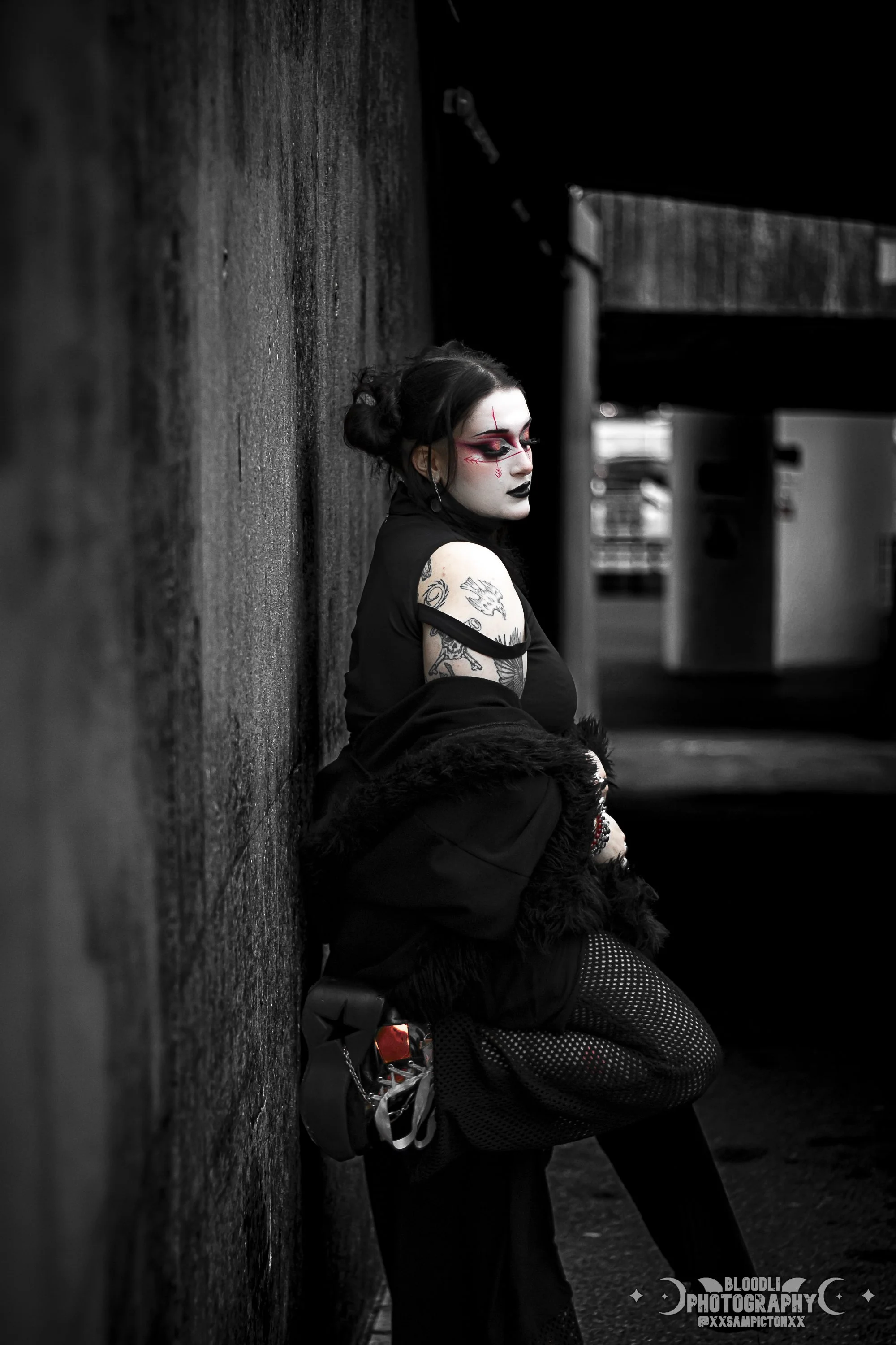 A woman with gothic makeup and tattoos leaning against a textured wall in an urban setting at night.