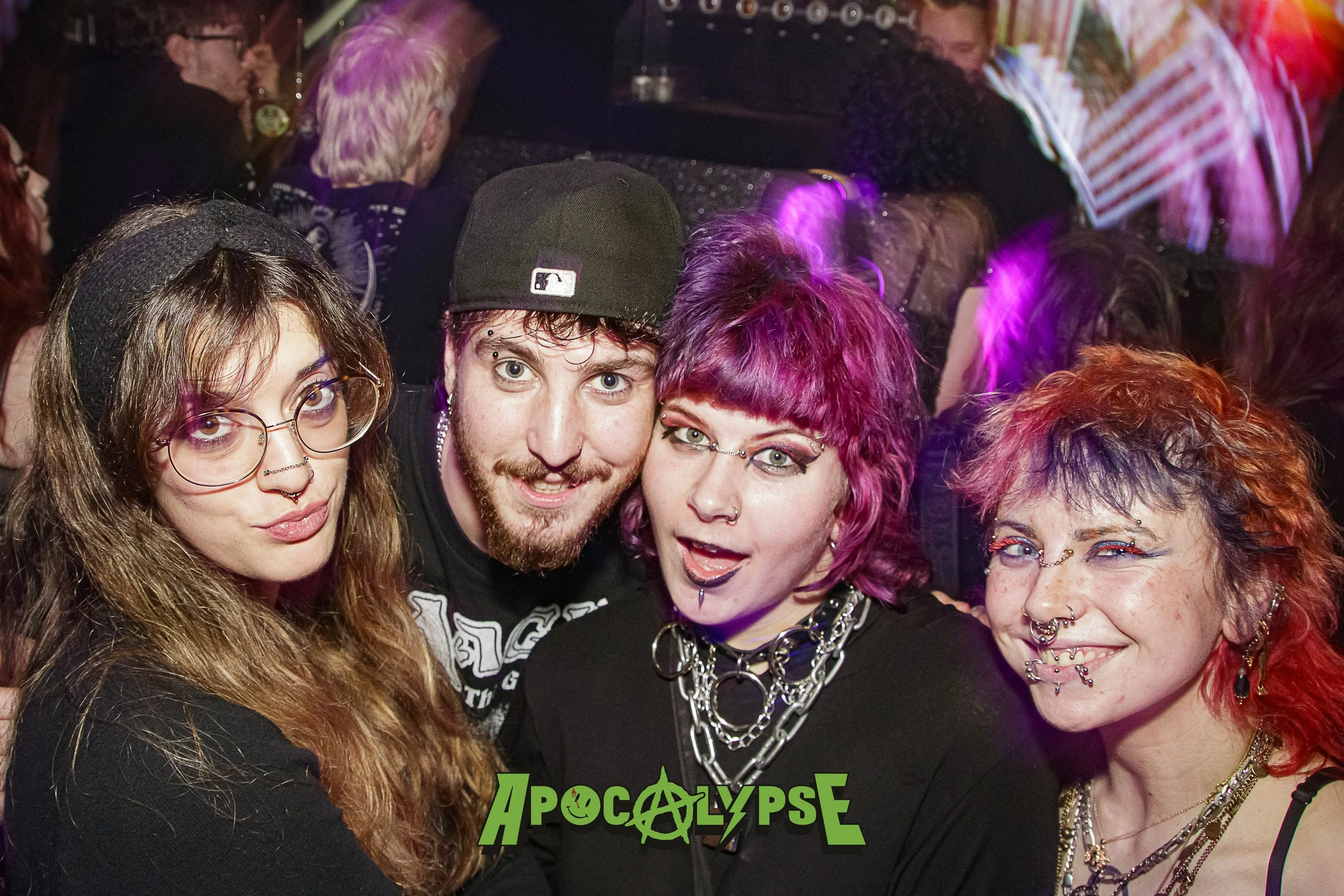 Group of four people with colorful hair and punk style accessories at a party with a crowded background and glowing lights.