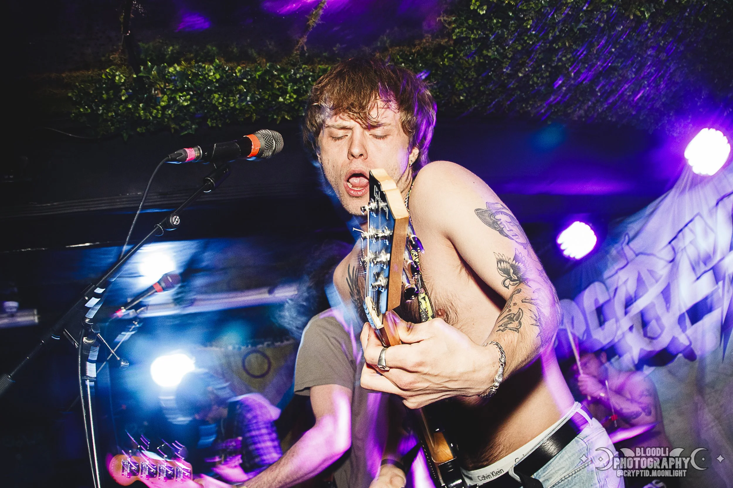 A shirtless male musician with tattoos playing an electric guitar on stage with colorful lights and a purple background.