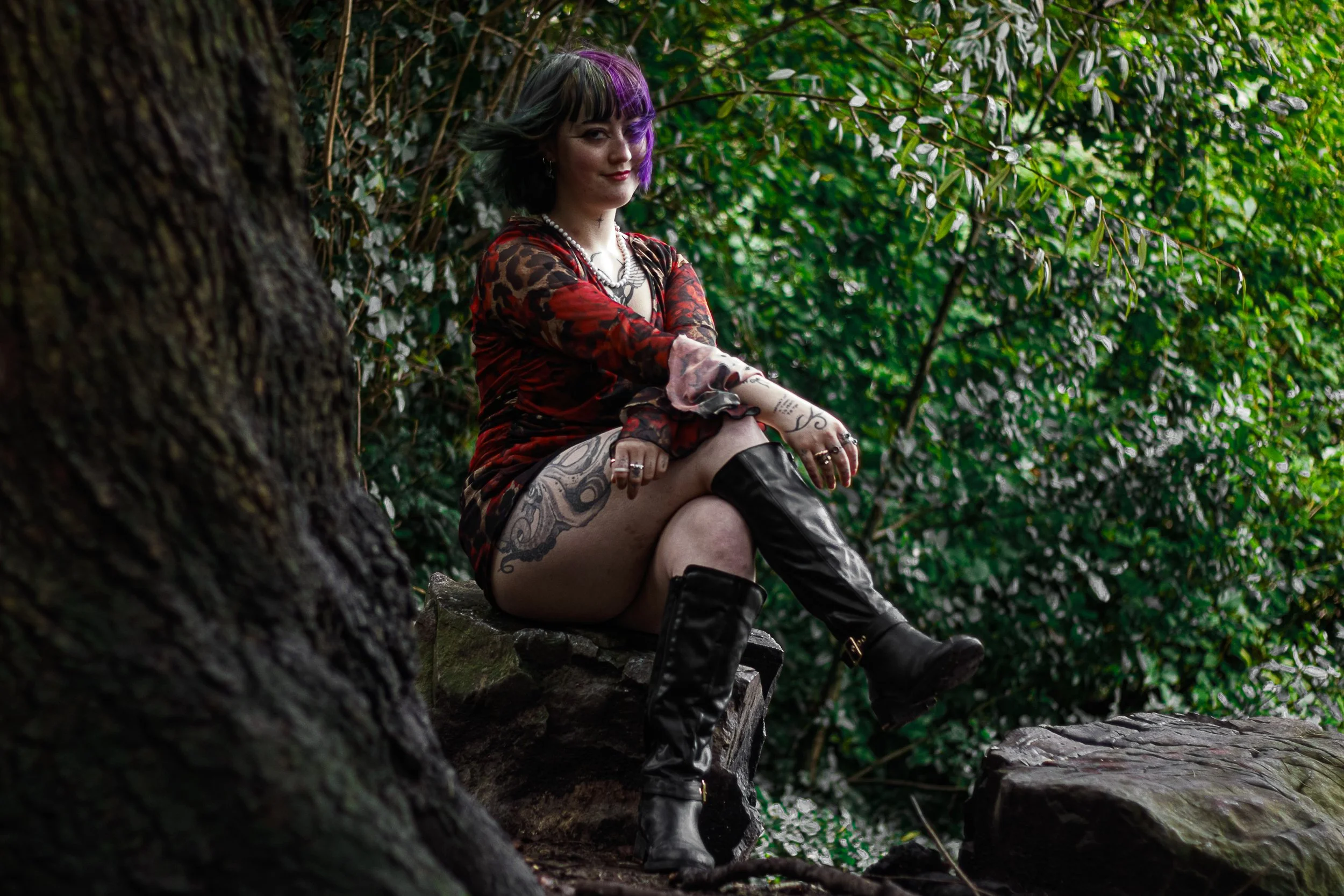 A woman with tattoos and purple hair sitting on a rock in a forest, wearing a red and black dress and black knee-high boots.
