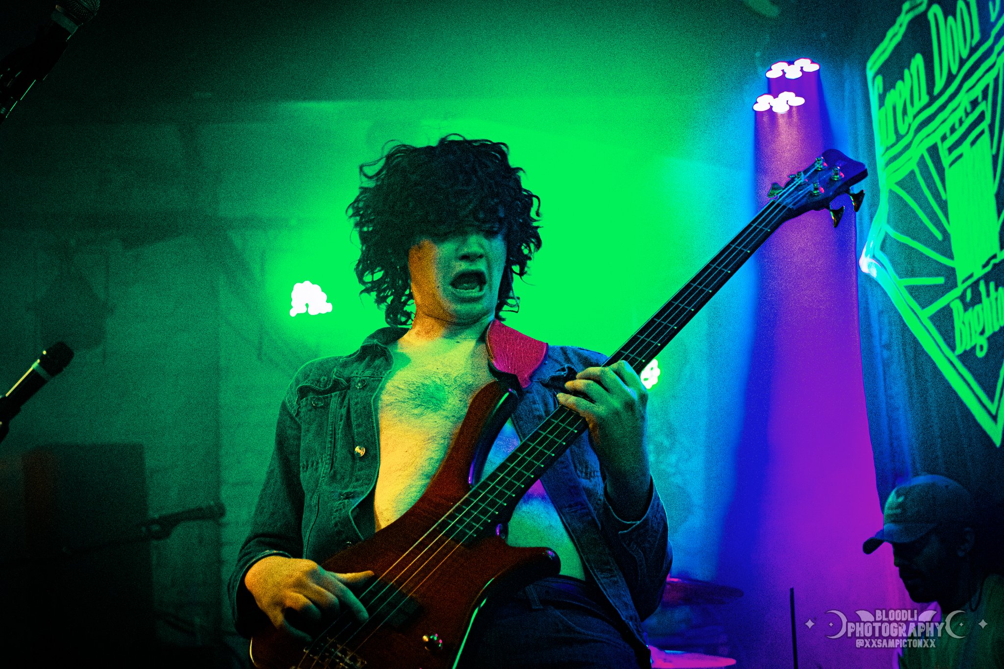 A person with curly hair playing an electric bass guitar on stage, with colorful lighting and a sign in the background.