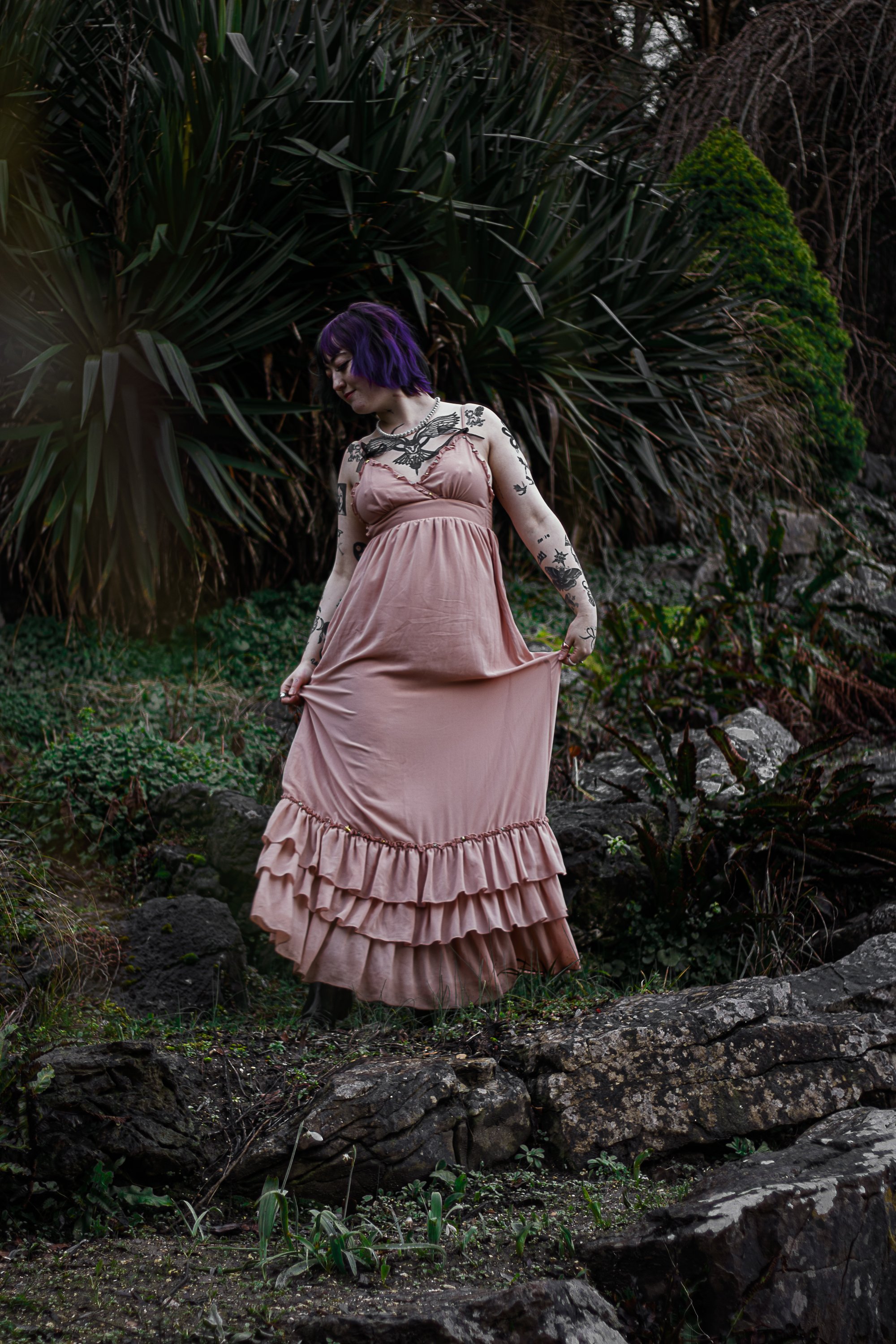 A woman with purple hair wearing a pink maxi dress with ruffles, standing outdoors on rocks surrounded by green plants and foliage.
