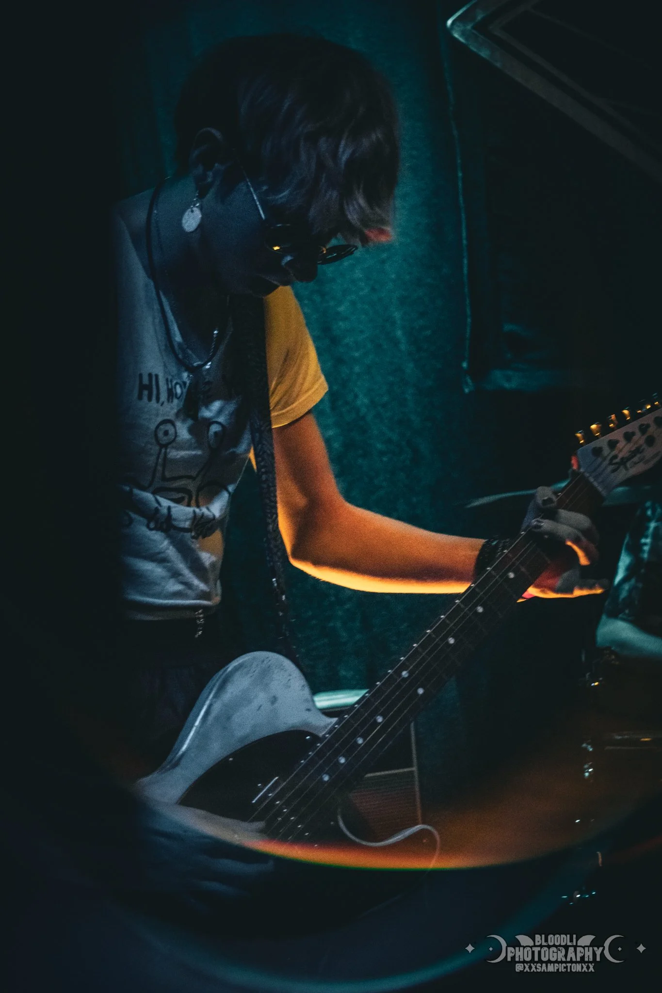 A person with short hair, wearing sunglasses and a graphic t-shirt, is playing an electric guitar in a dark room.
