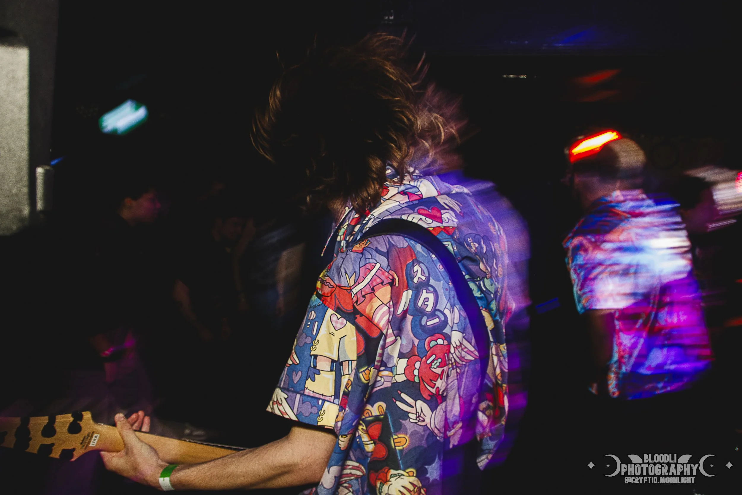 Person playing electric guitar at a dark, crowded concert or party with blurred colorful lights and other people in the background.