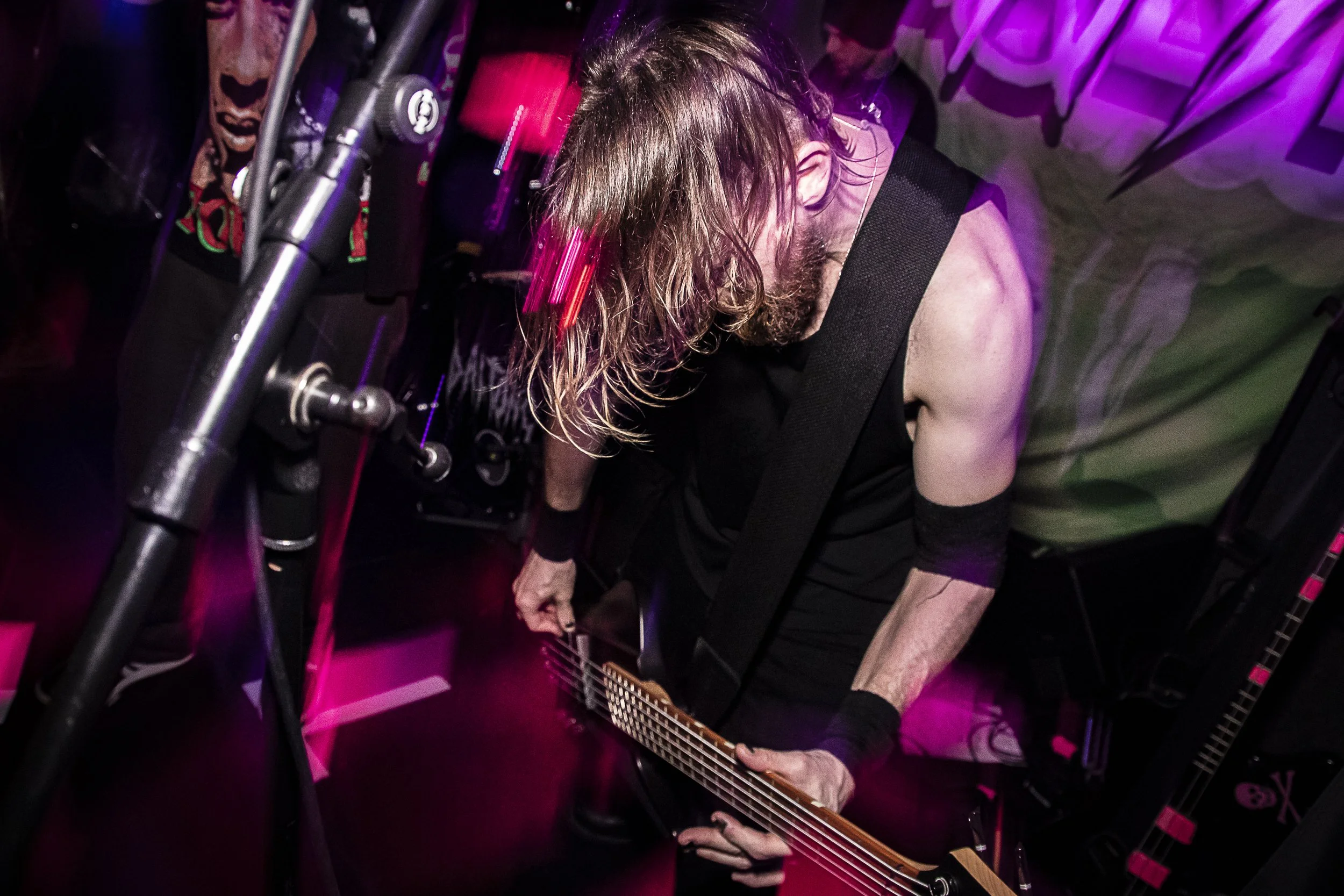 A musician playing an electric guitar on stage with pink and purple lighting.