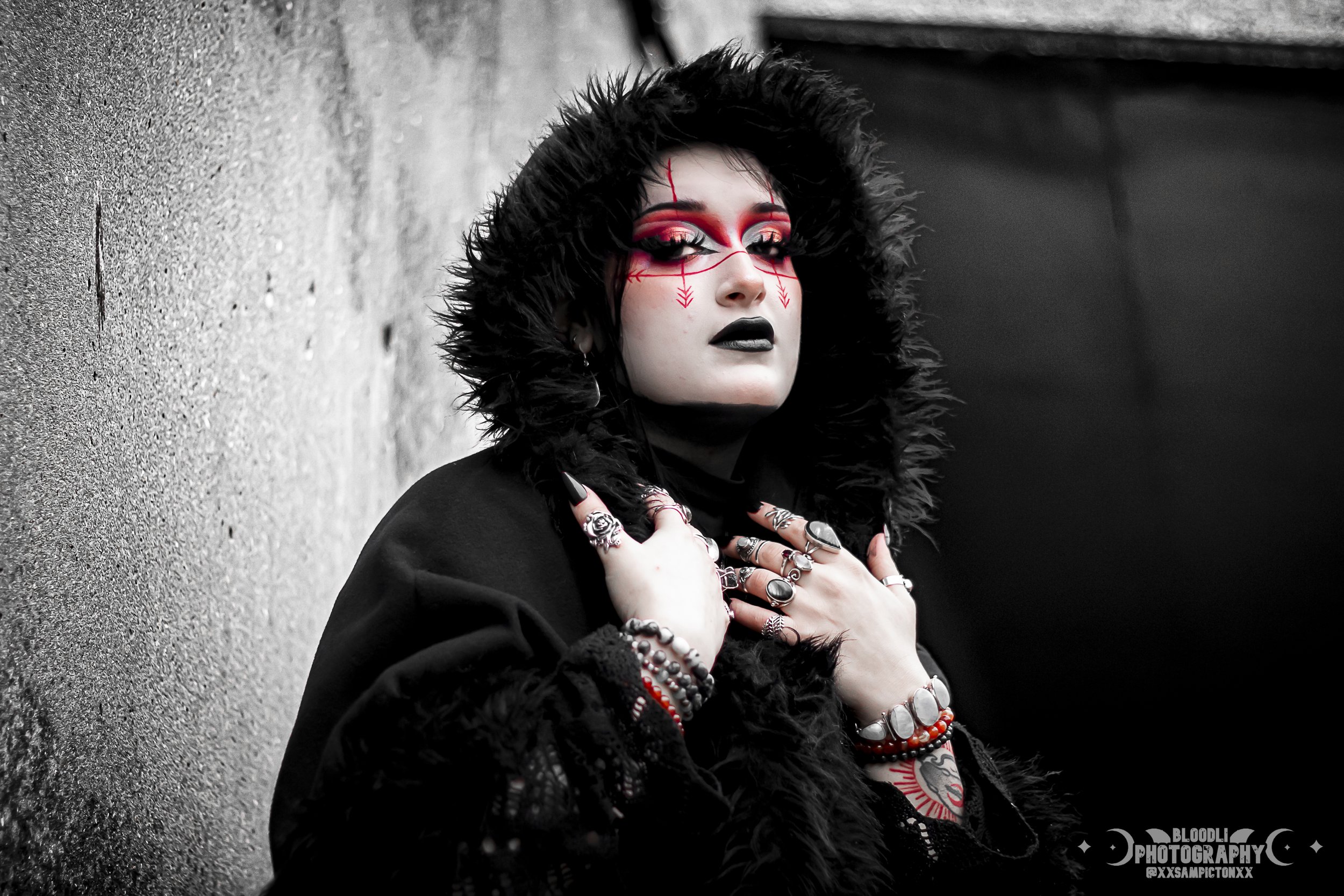 A person with pale skin and dark makeup, dressed in black with a furry hood, standing against a gray wall. They wear multiple rings, bracelets, and necklaces, with eye-catching red and black face paint and makeup. Their expression is intense and conf