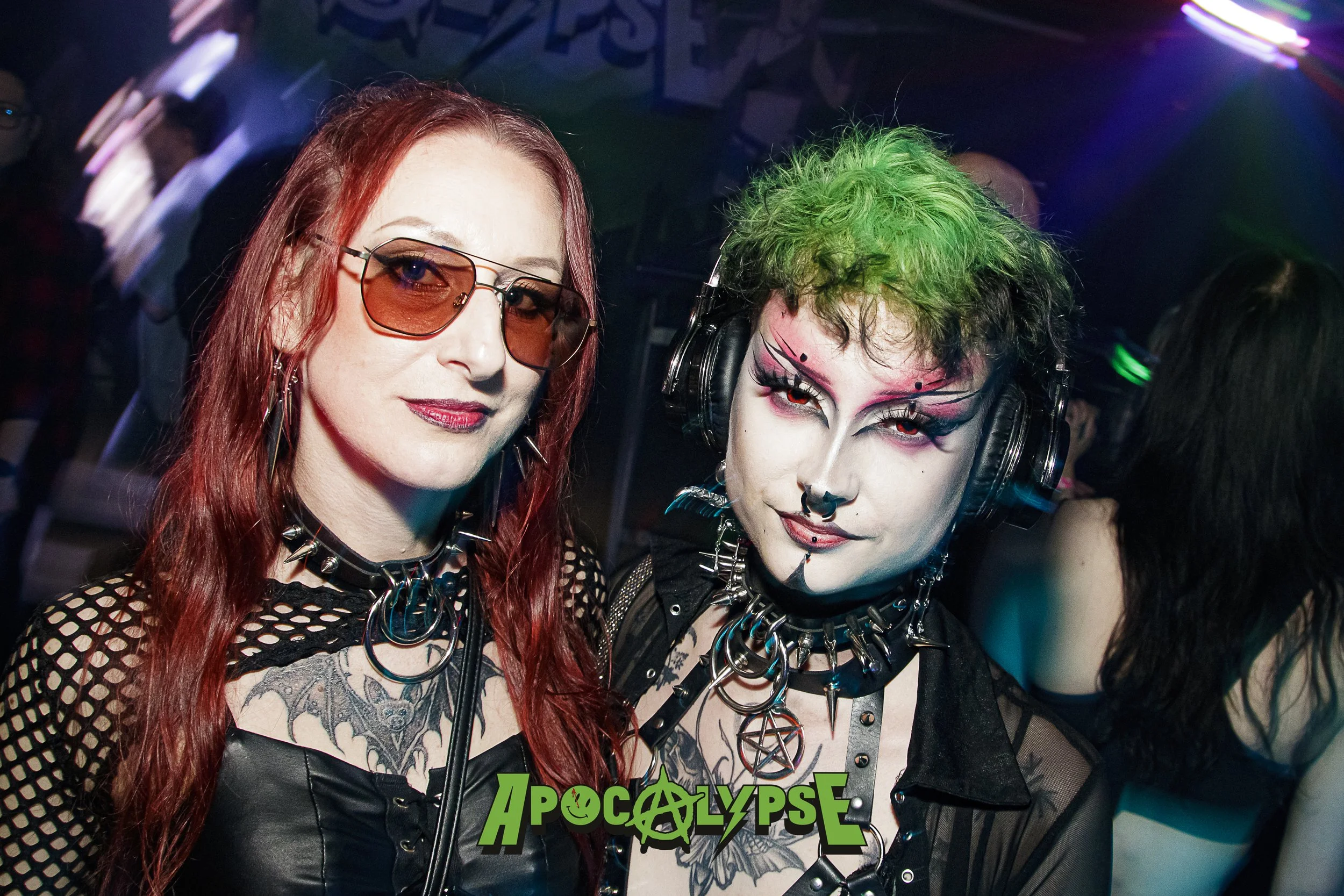 Two women dressed in goth style at a party or club, with one woman having red hair, wearing orange sunglasses, and a fishnet top, and the other woman with green hair, face makeup, and various piercings, and wearing dark clothing. The background has c