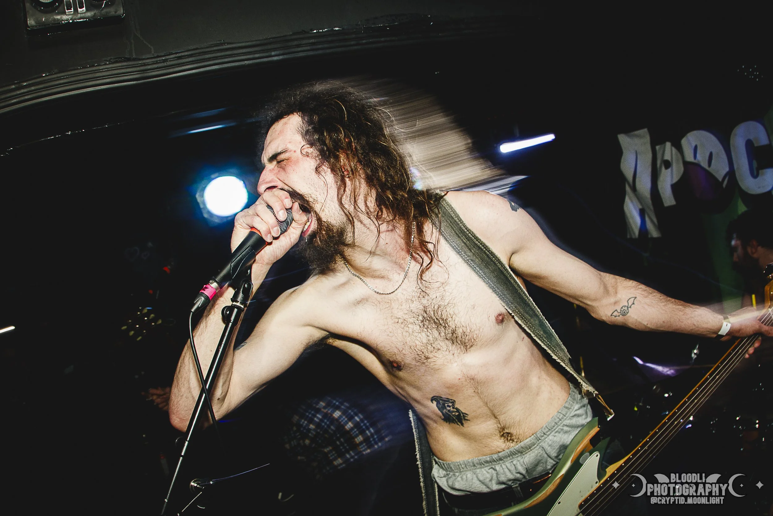 A shirtless man with long hair and tattoos, singing into a microphone on stage with colored lights and a dark background.