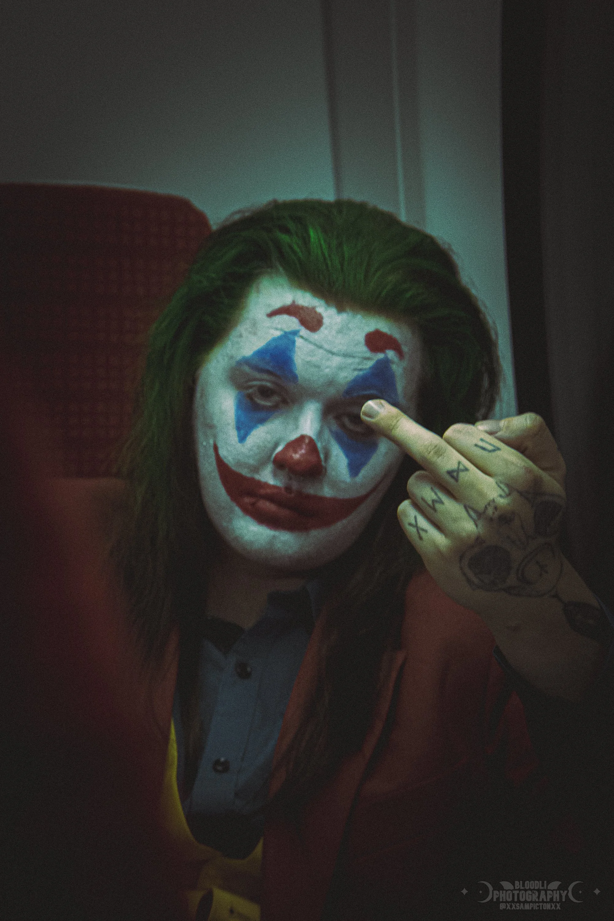 Person with clown makeup, green hair, and tattoos on hand, making a rude gesture