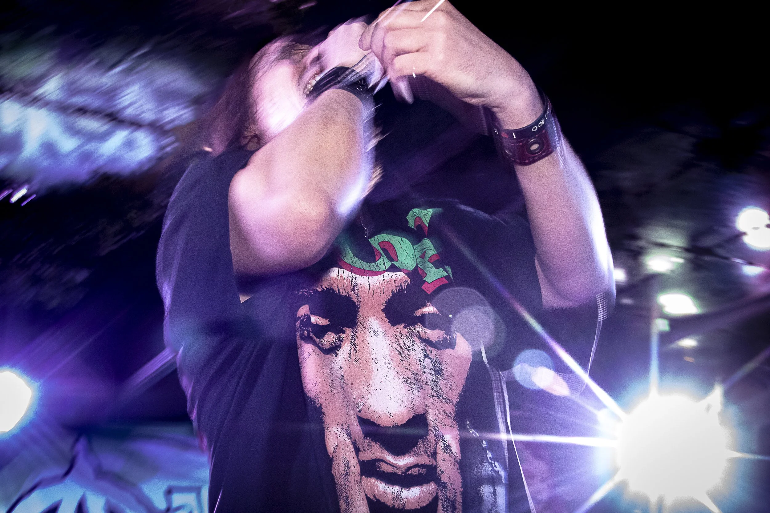 A person performing on stage with a microphone, wearing a black t-shirt with a graphic of a woman's face on it, surrounded by bright stage lights.