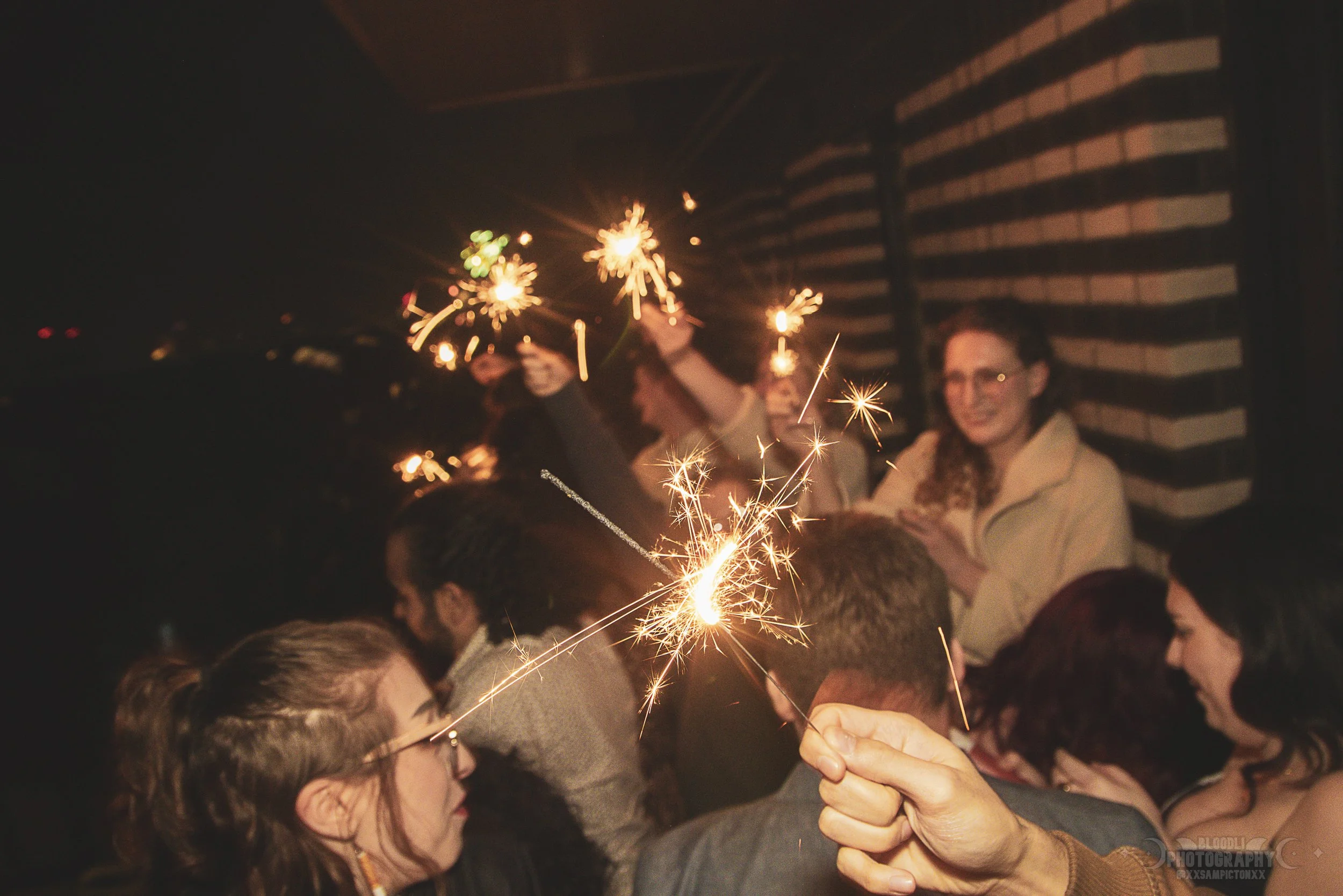 Group of people celebrating with sparklers at night, some smiling and enjoying the moment.