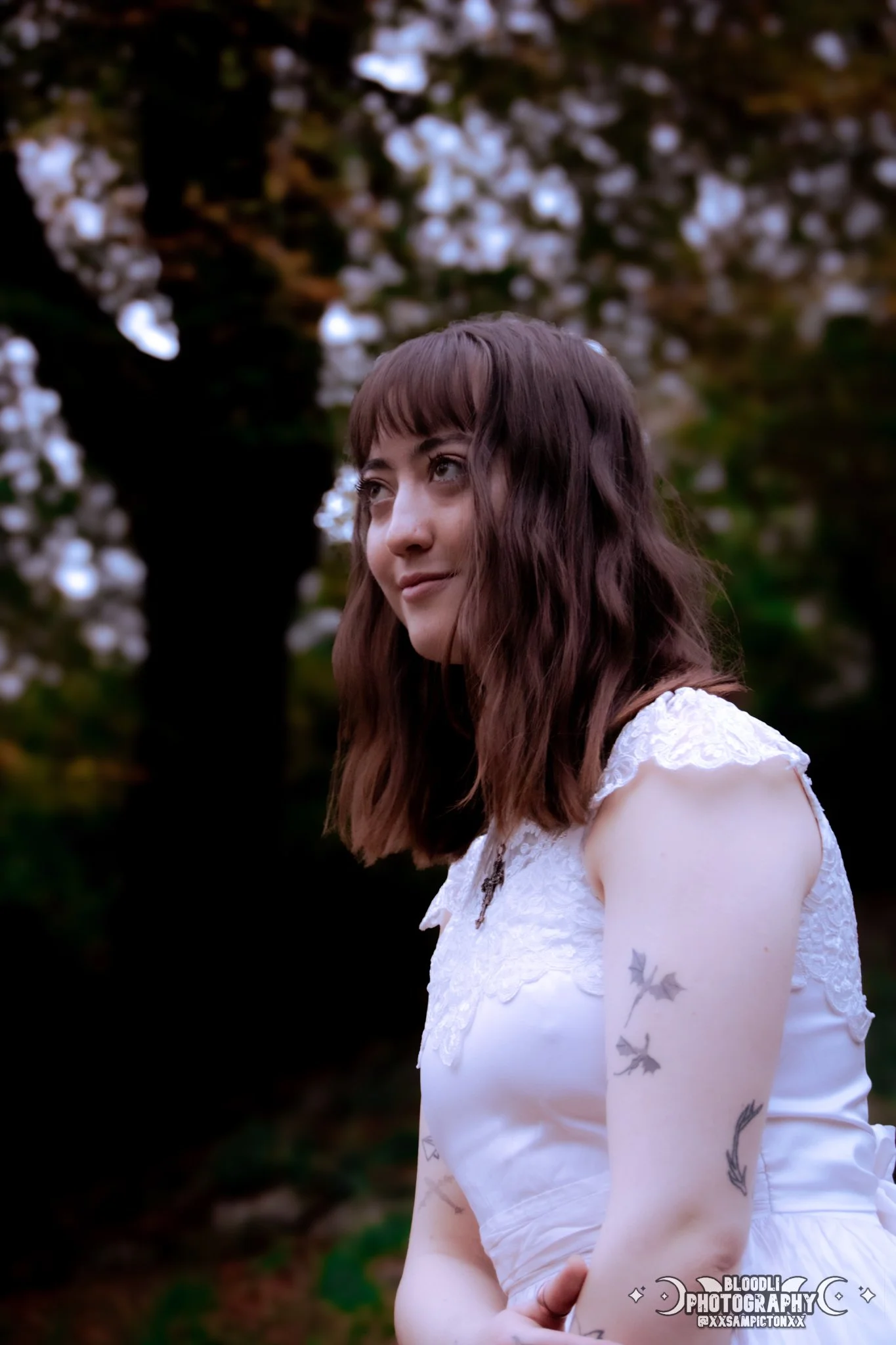 A young woman with shoulder-length wavy brown hair and tattoos on her left arm, wearing a white lace dress with a key necklace, standing outdoors in front of a blurred background with blooming white flowers and dark trees.