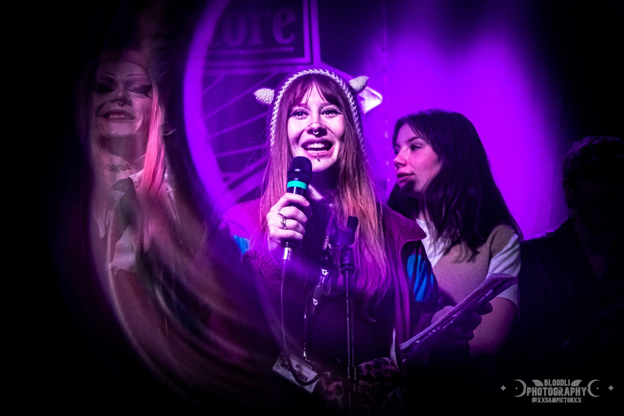A young woman with long red hair, wearing a sheep headband and a purple jacket, speaking into a microphone at a dimly lit event with purple lighting.