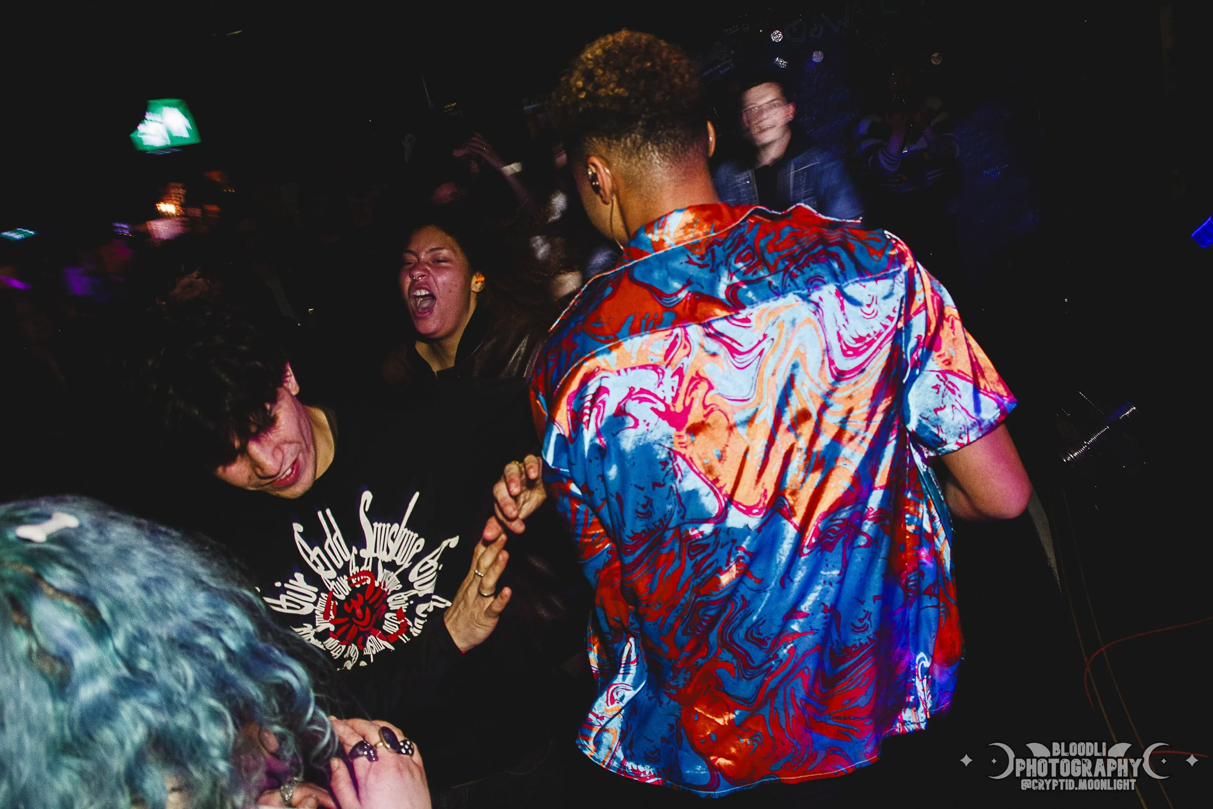 People dancing and enjoying music at a party, with vibrant clothing and energetic expressions, in a dimly lit nightclub or concert setting.