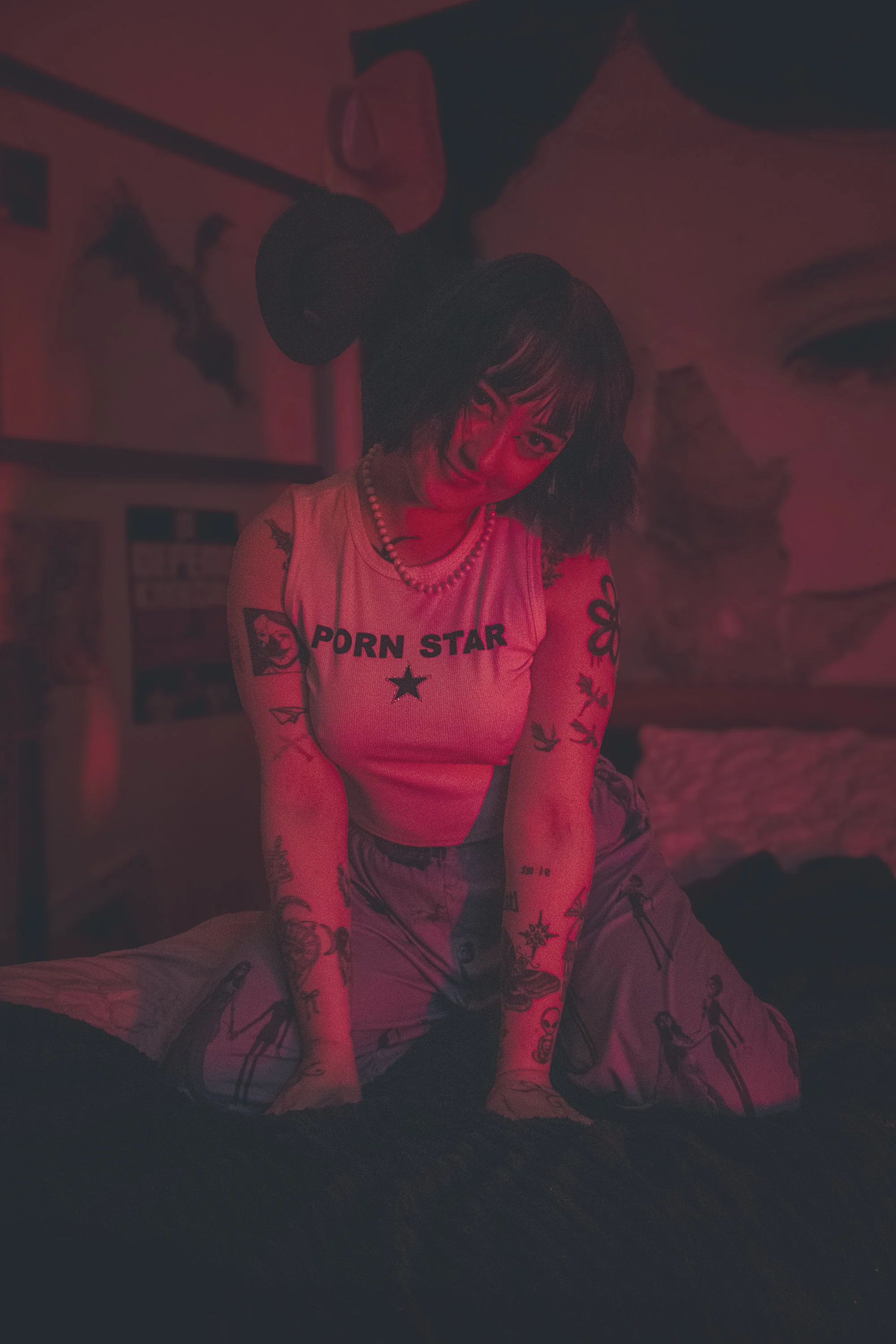 A woman with tattoos and short dark hair posing on her knees on a bed in a dimly lit room with red lighting, wearing a sleeveless shirt that says 'PORN STAR', patterned pants, and a beaded necklace.