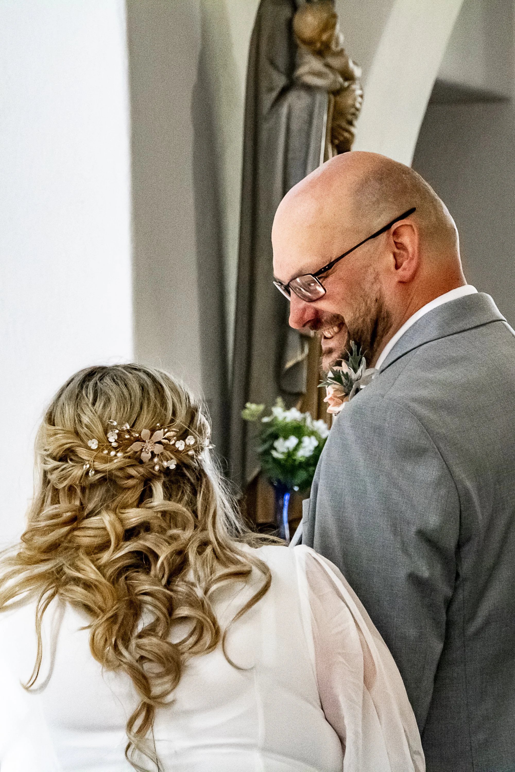 A bald man with glasses smiling at a woman with long, curly blonde hair wearing a floral hairpiece at a wedding ceremony.