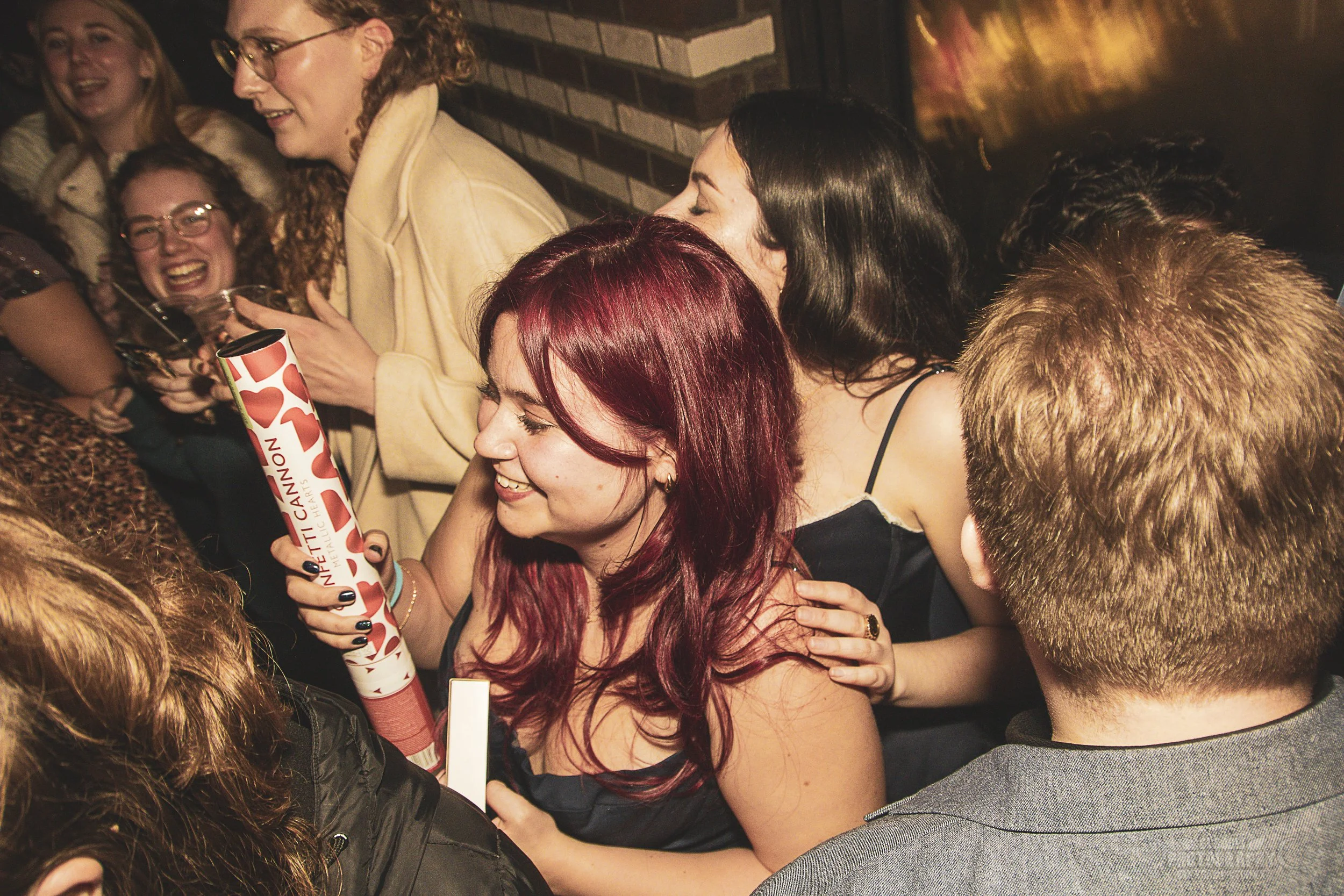 A group of people celebrating together, with a woman holding a cylindrical gift box, all smiling and laughing in a lively atmosphere.