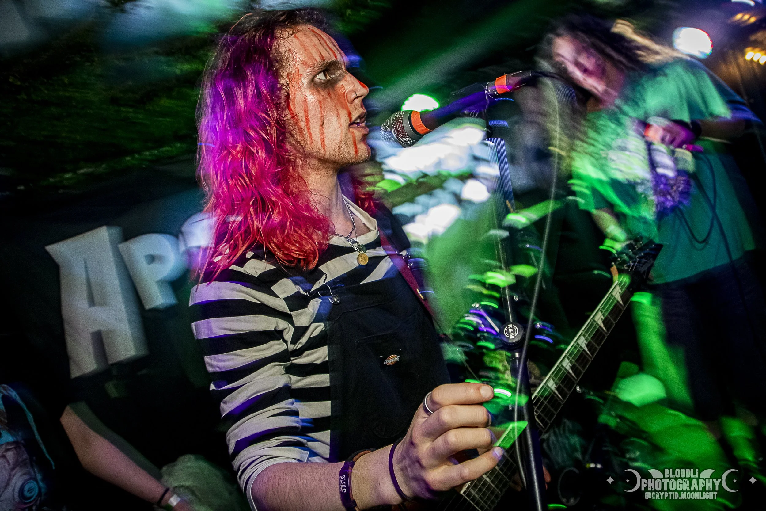 A musician with vibrant pink hair and striped shirt performing at a concert, with blurred colorful lights and a guitar in hand.