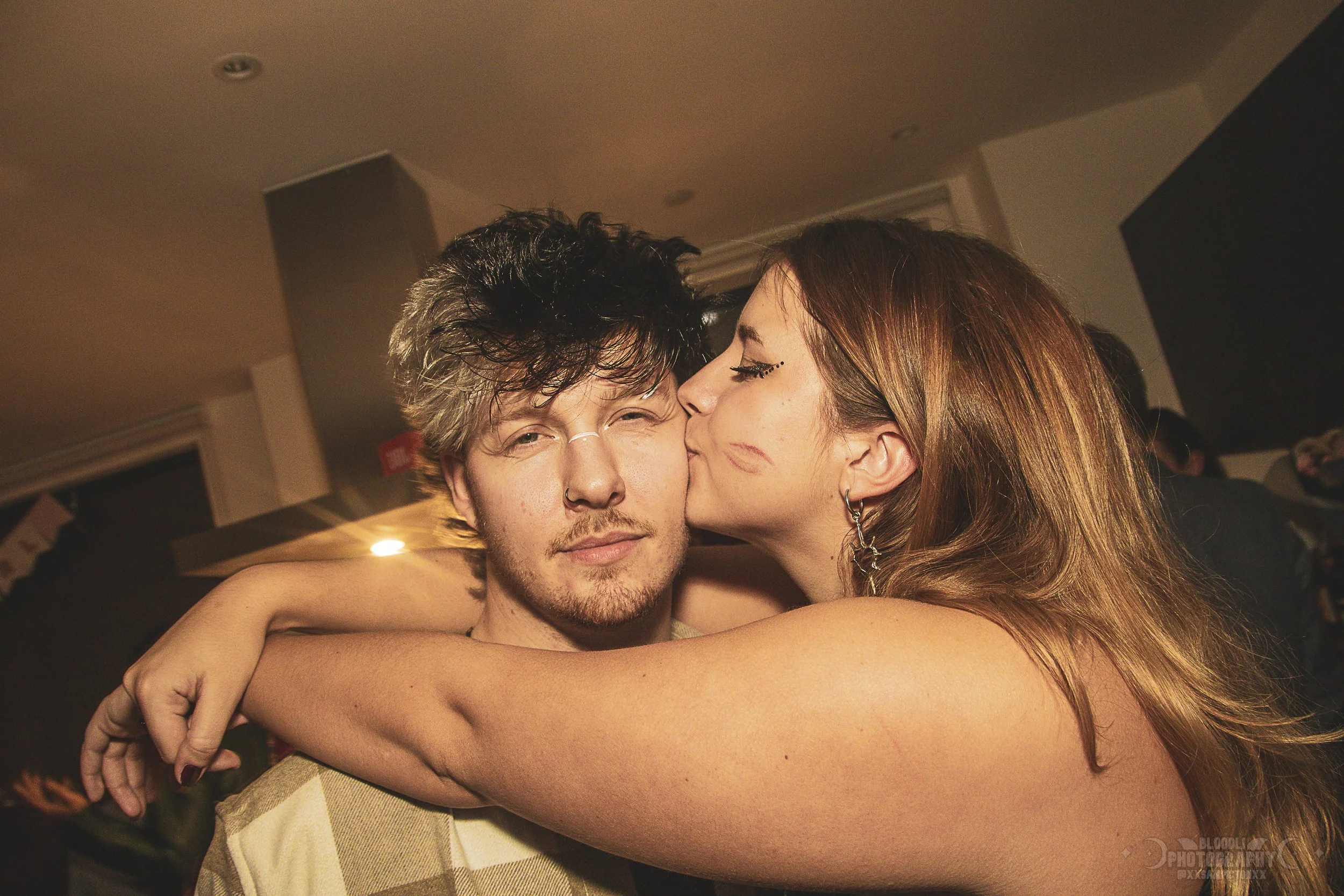 A young woman is kissing a young man on the cheek at a party or social gathering. The man has tousled dark hair, facial hair, and is making a winking face, while the woman has long, wavy reddish-brown hair and earrings.
