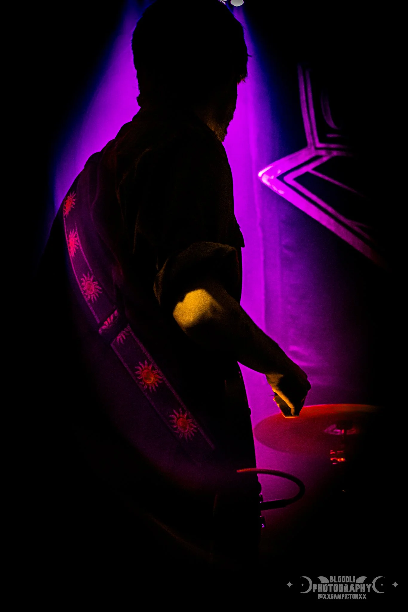 Silhouette of a person with curly hair with purple and pink neon lighting, standing next to a table with a disc, possibly in a nightclub or concert setting.