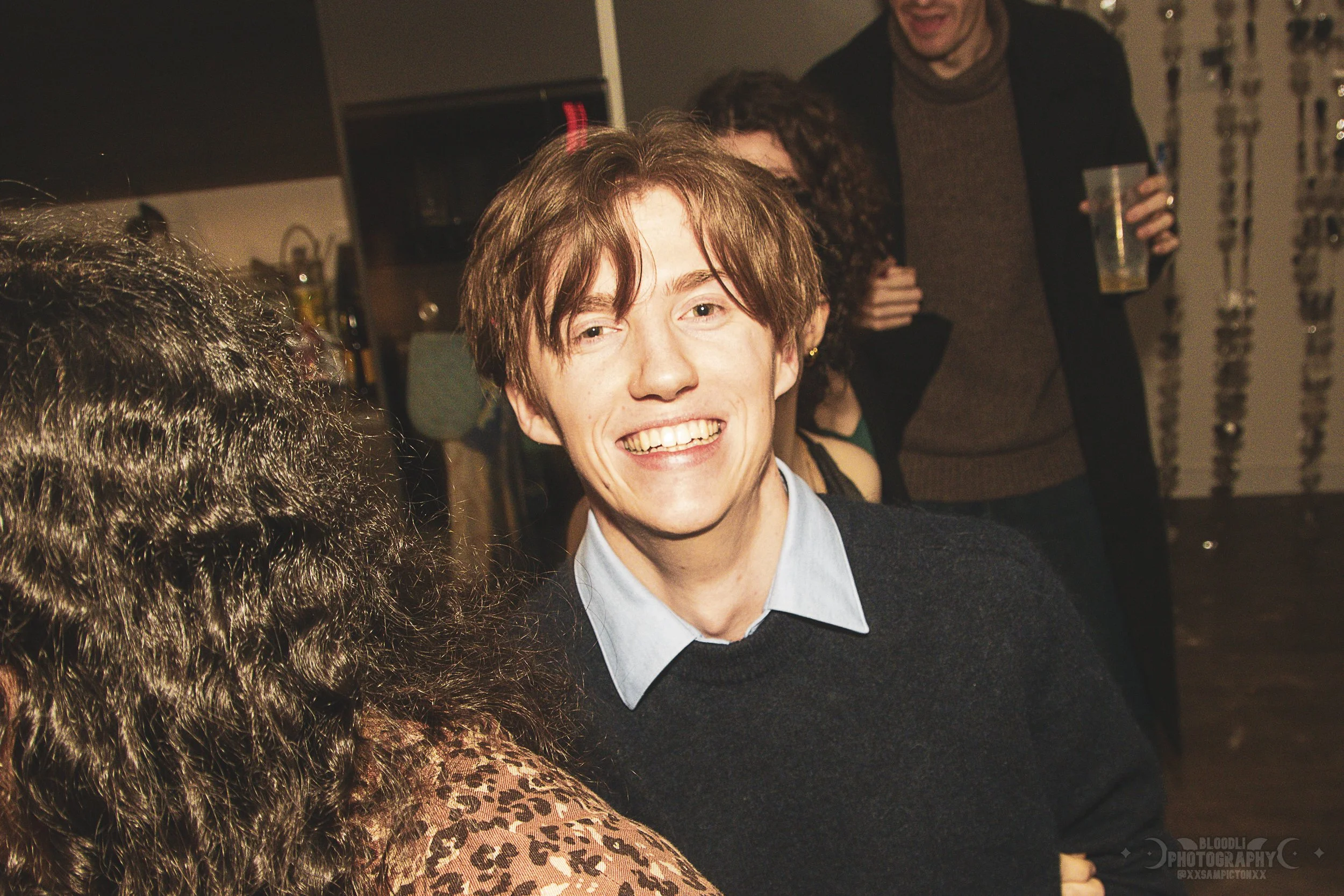 A young man with brown hair, wearing a dark sweater over a light blue shirt, smiling at a social gathering with people in the background holding drinks.