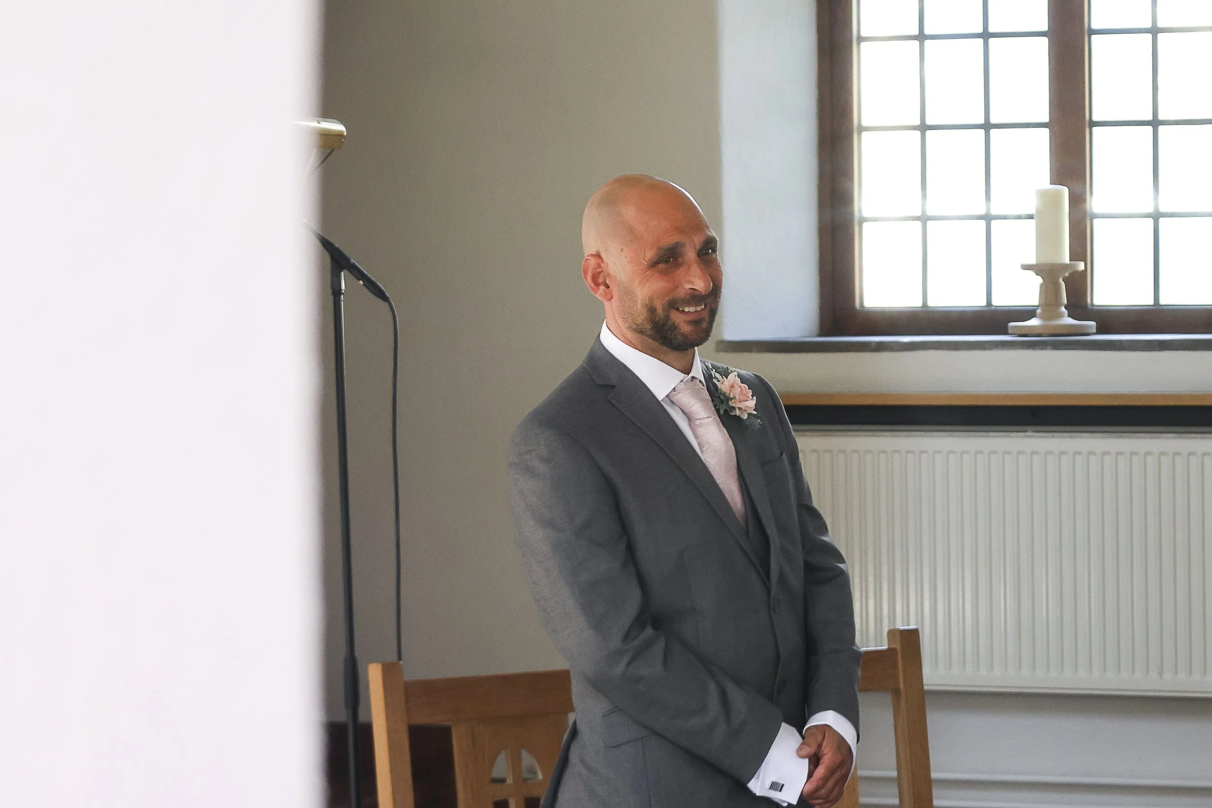 A man dressed in a gray suit with a white shirt and pink tie, wearing a pink flower boutonniere, standing indoors near a large window with multiple panes, smiling.