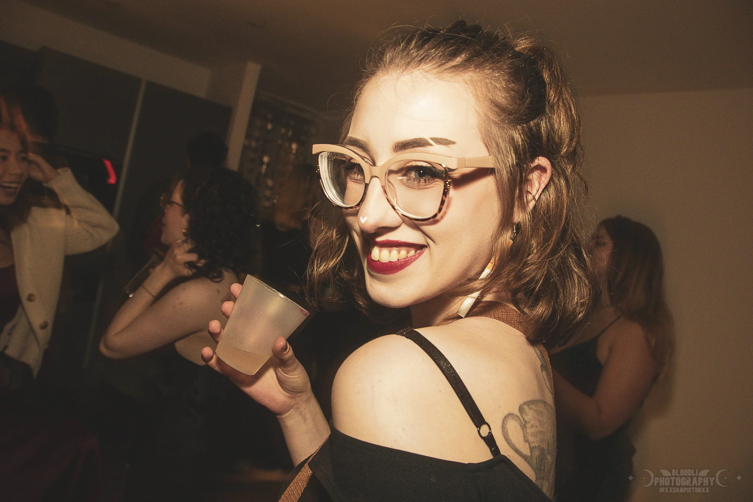 A young woman with glasses, a tattoo on her shoulder, and wavy hair at a party, smiling while holding a drink. Other people are in the background.