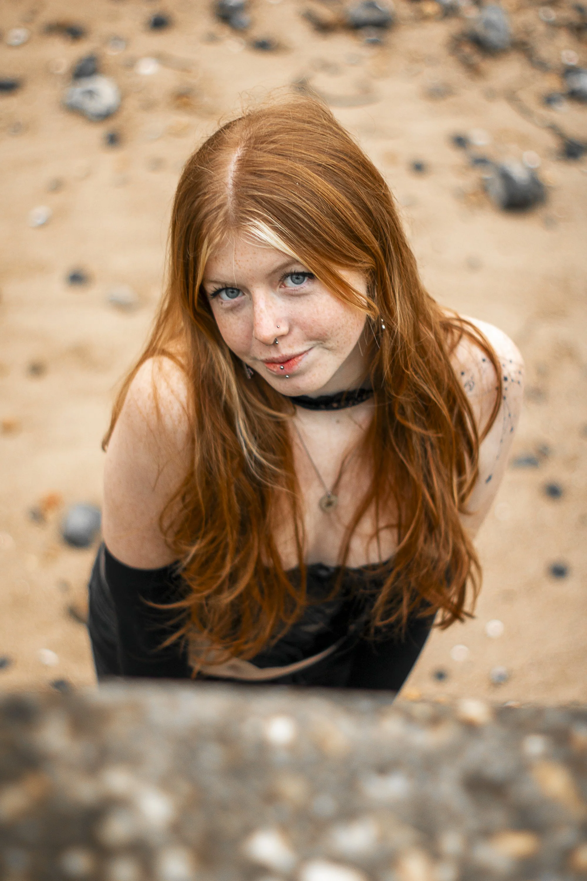 A young woman with long red hair and blue eyes, looking up and smiling at the camera on a sandy beach with scattered rocks.