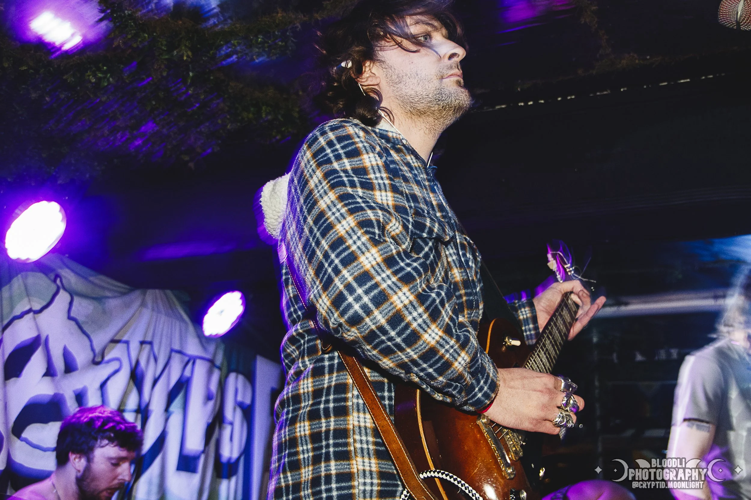 A male musician playing an electric guitar during a live performance, wearing a plaid shirt, with purple stage lights and a band banner in the background.