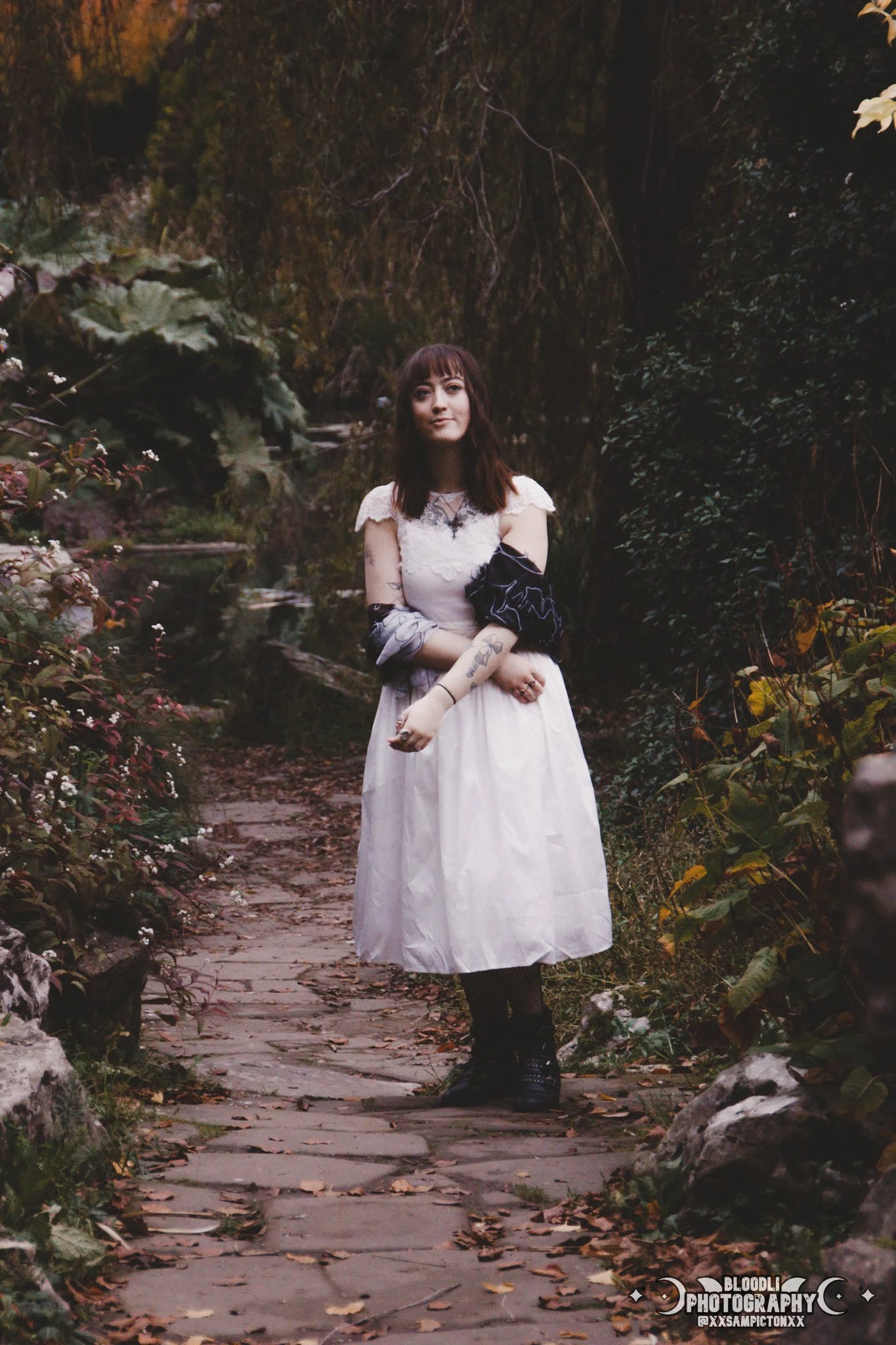 A young woman with dark brown hair and tattoos standing on a stone path surrounded by greenery, wearing a white dress and black boots, in a forest setting.