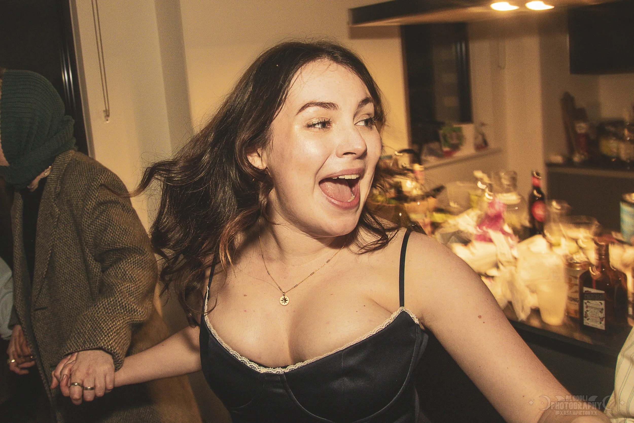 Young woman with long dark hair in a black camisole with lace, smiling and holding hands with someone at a party, with a cluttered kitchen in the background.